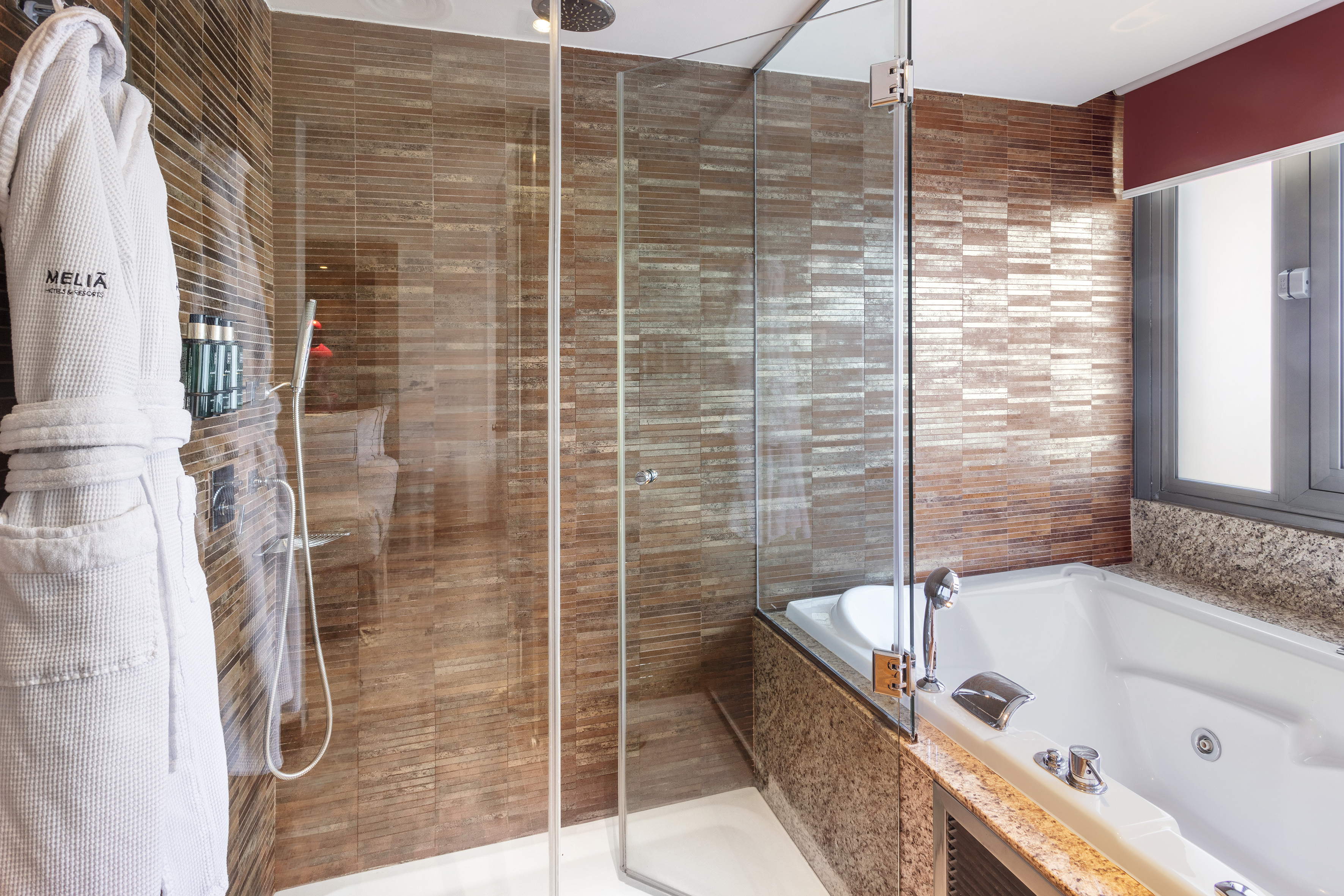 Baño moderno de hotel con ducha de cristal, bañera, azulejos marrones y albornoz Meliã.