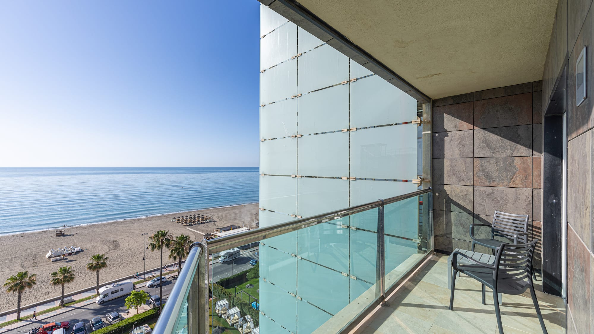 Sunny hotel balcony with ocean view, sandy beach, and palm trees.