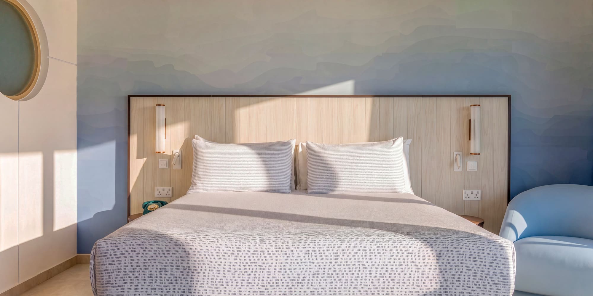 Modern hotel room, large bed, blue gradient wall, wood headboard, sunlit.