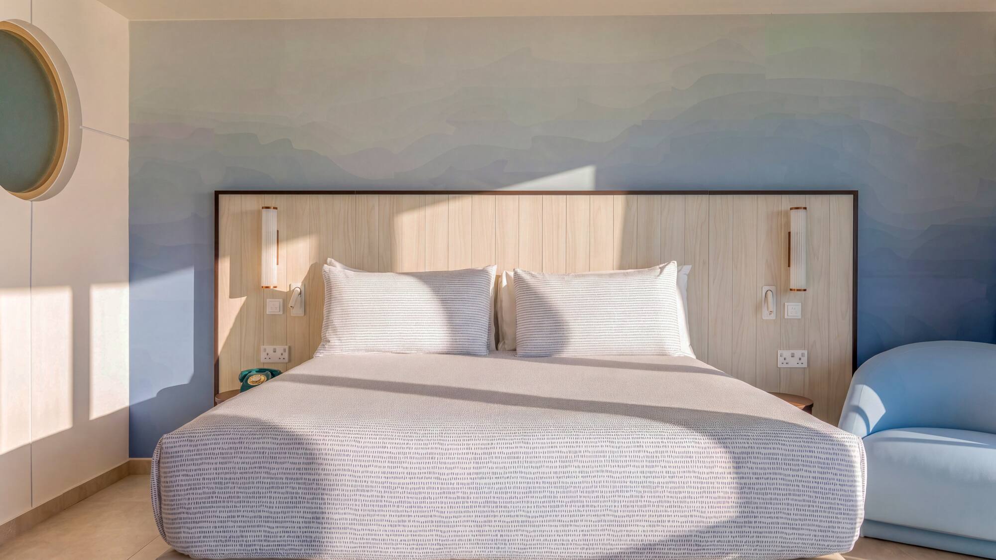 Modern hotel room, large bed, blue gradient wall, wood headboard, sunlit.