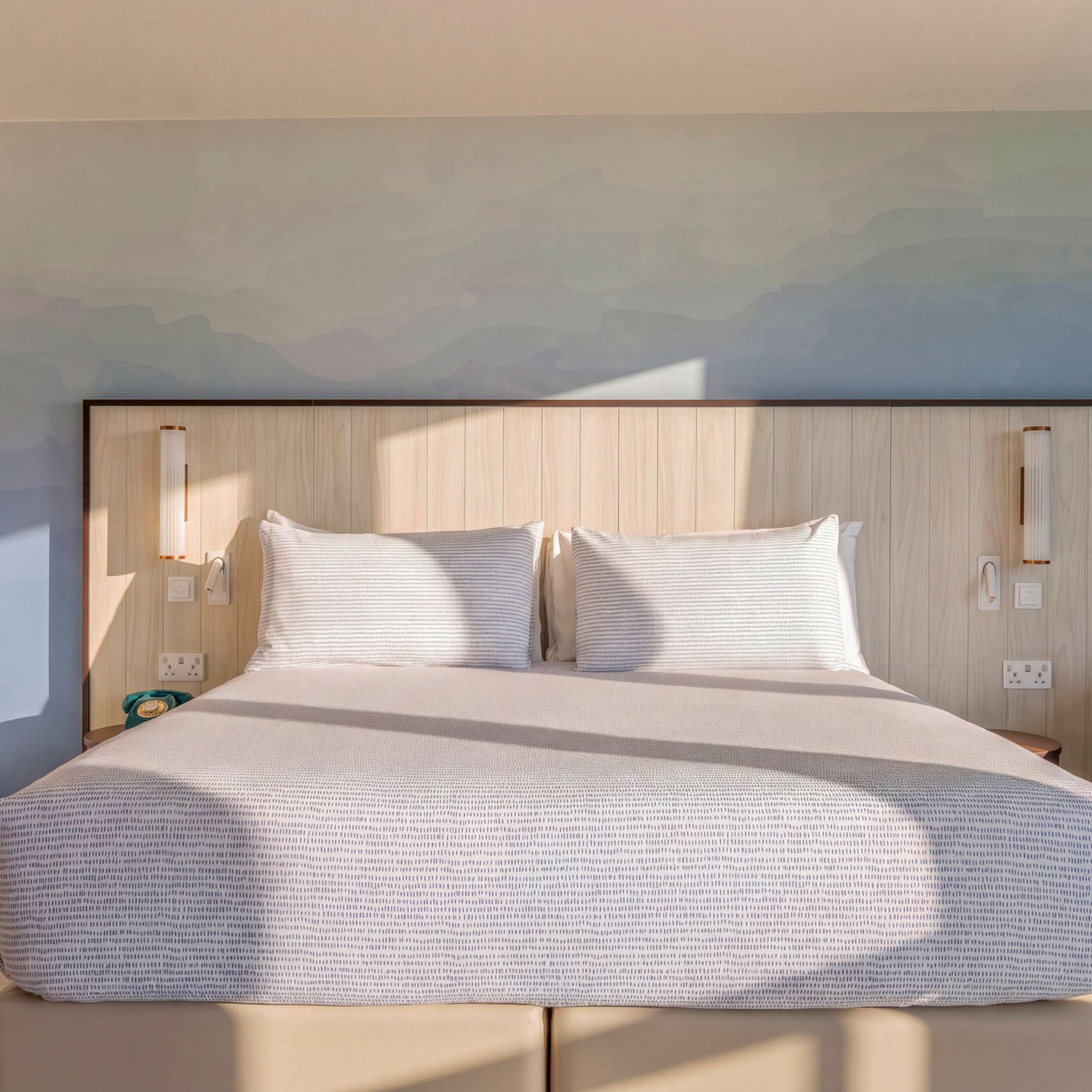 Modern hotel room, large bed, blue gradient wall, wood headboard, sunlit.