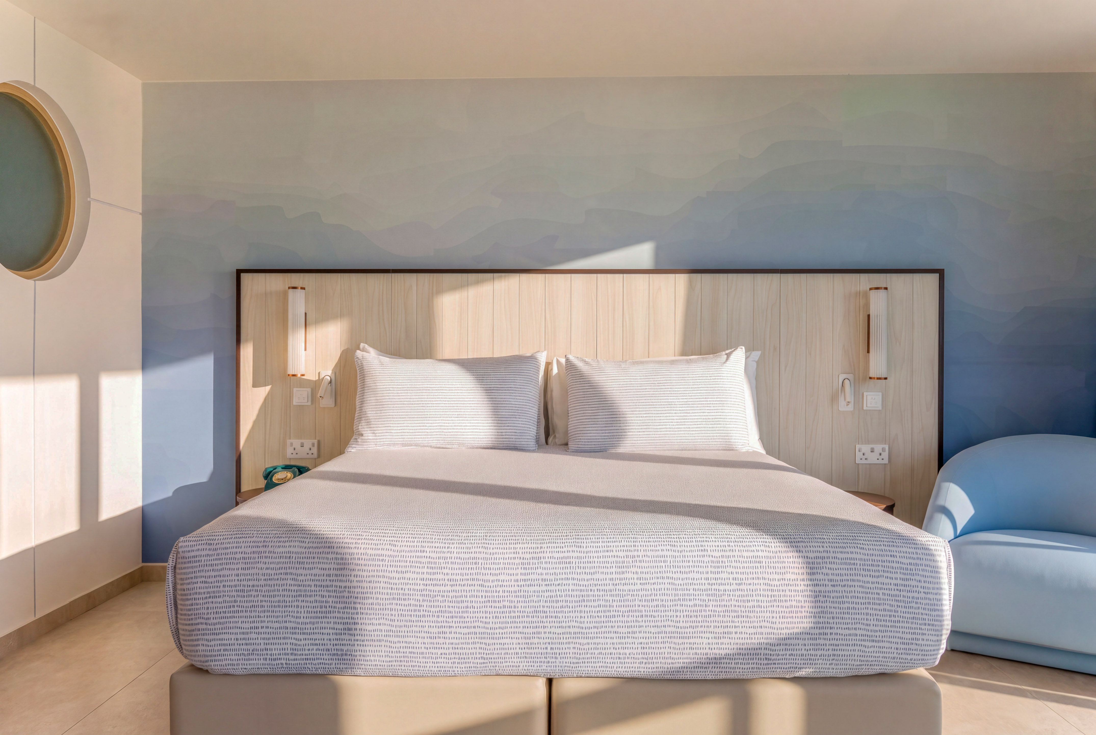 Modern hotel room, large bed, blue gradient wall, wood headboard, sunlit.