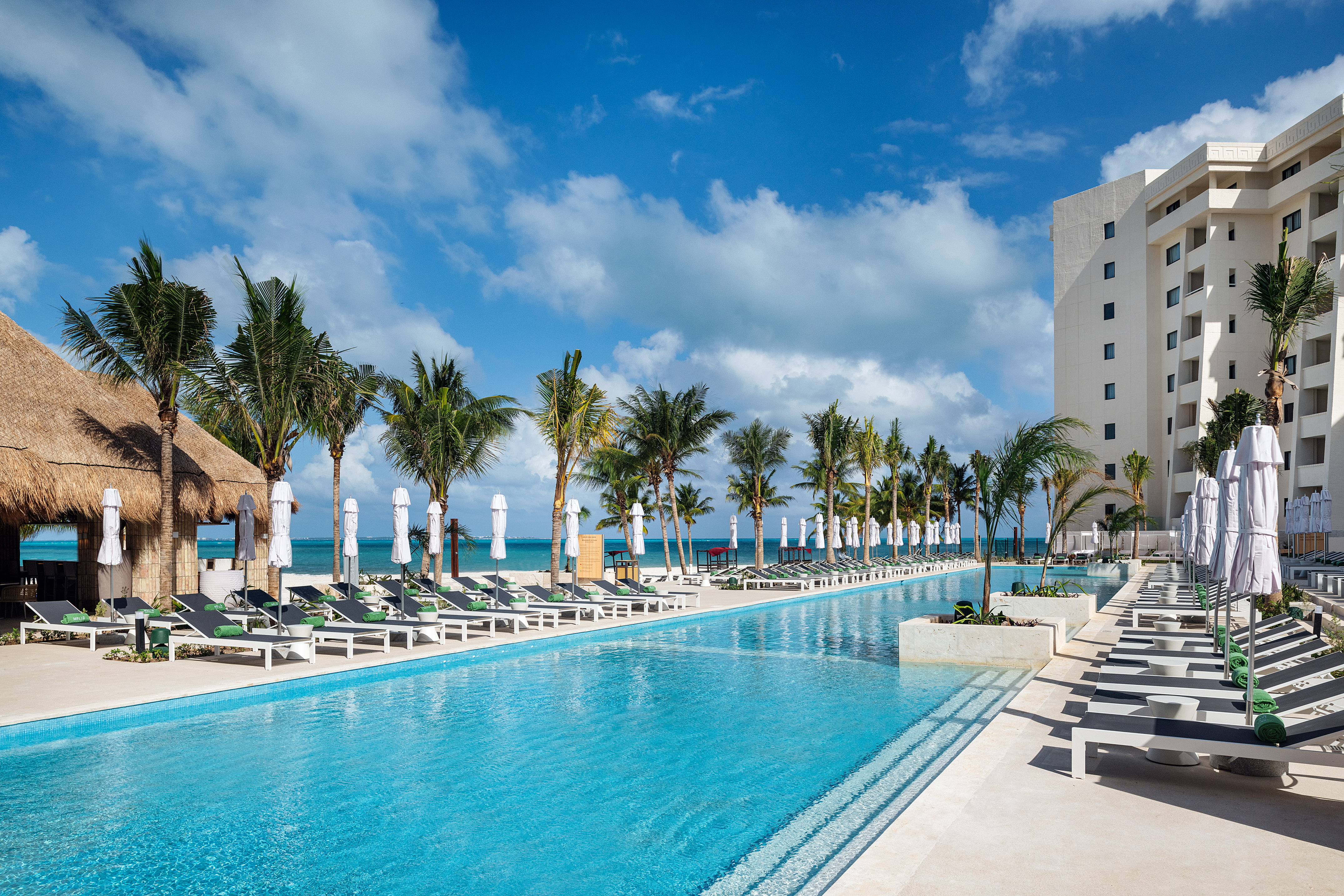 Tropical resort pool with palm trees, sun loungers, and hotel building.
