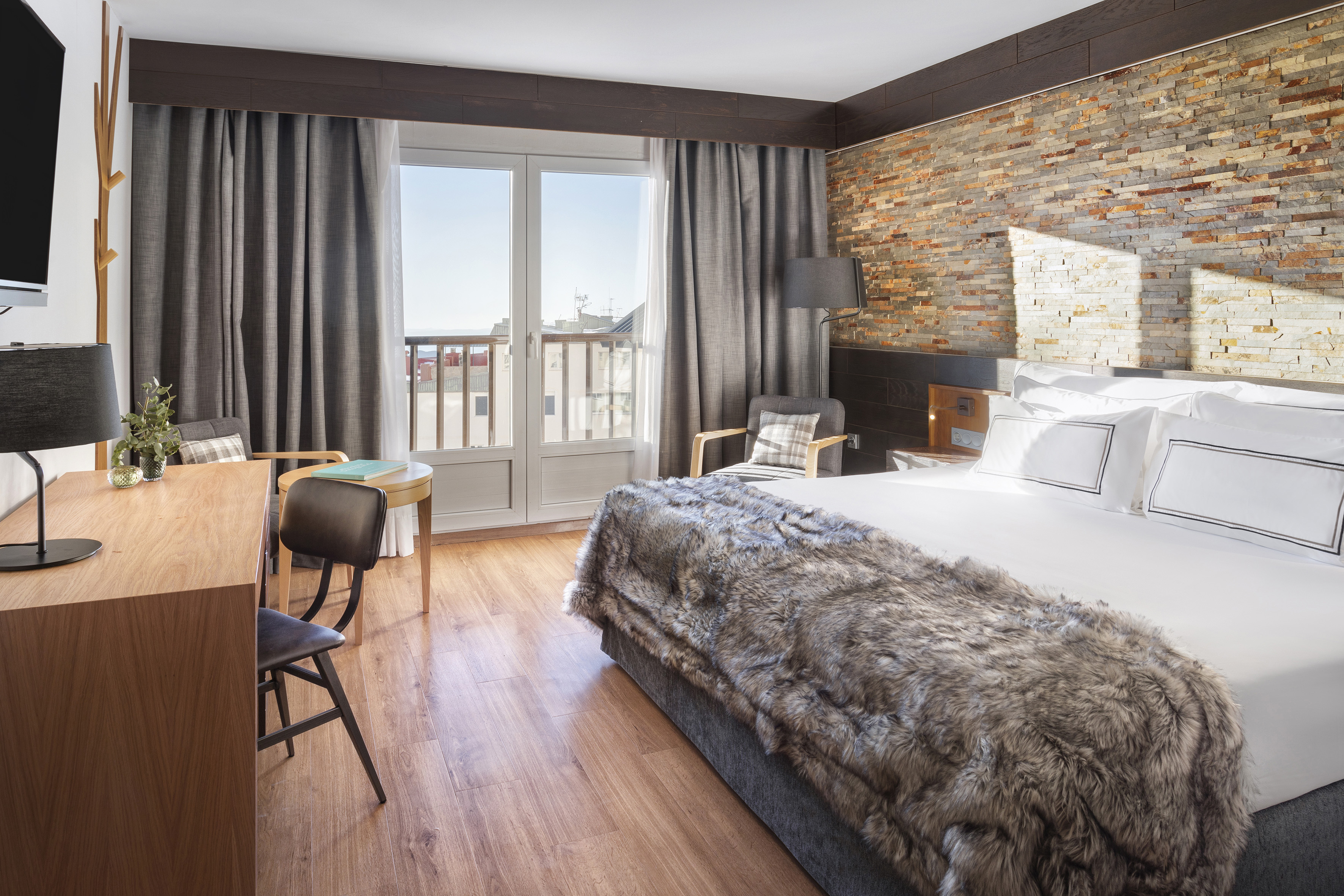 Modern hotel room with large bed, faux fur throw, stone wall, wood floor, balcony.