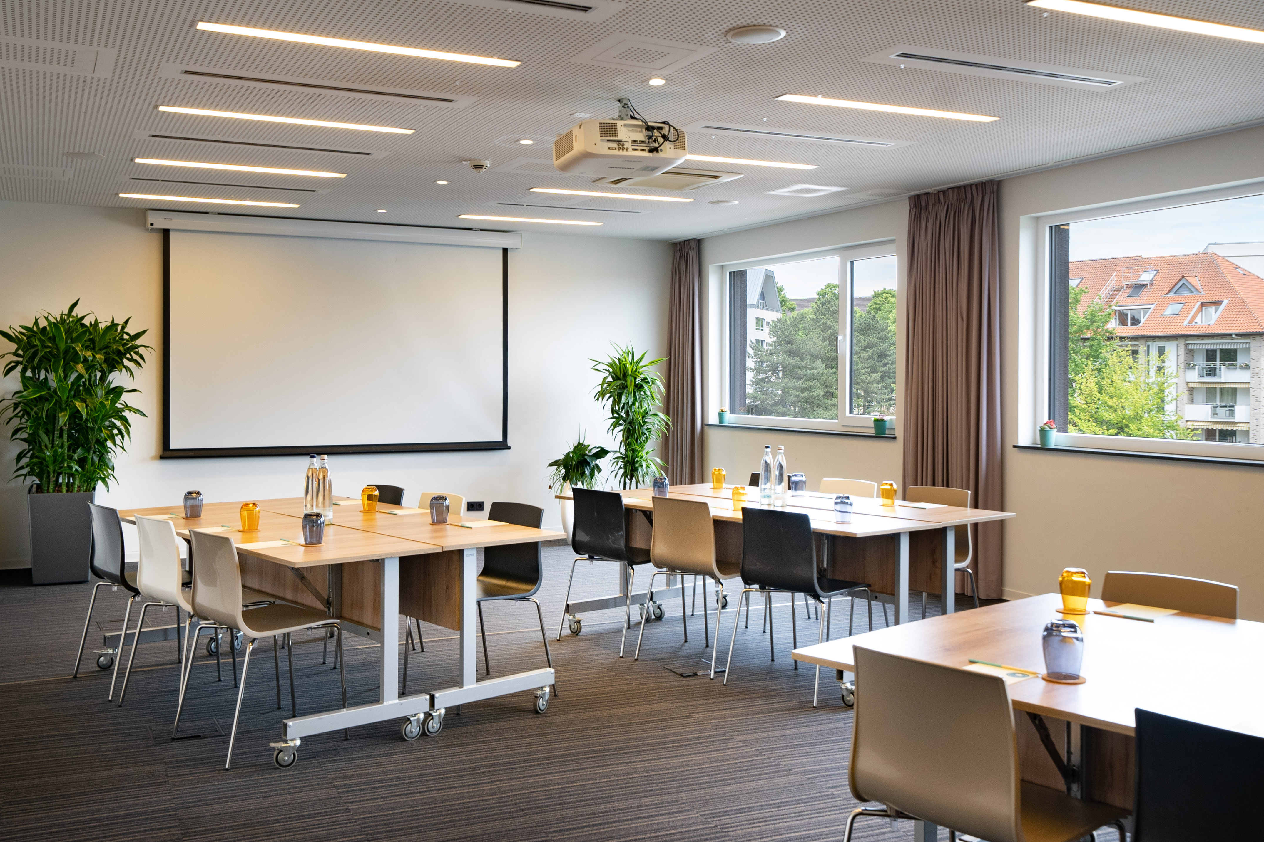 Spacious meeting room with flexible tables, chairs, projector, screen, and plants.
