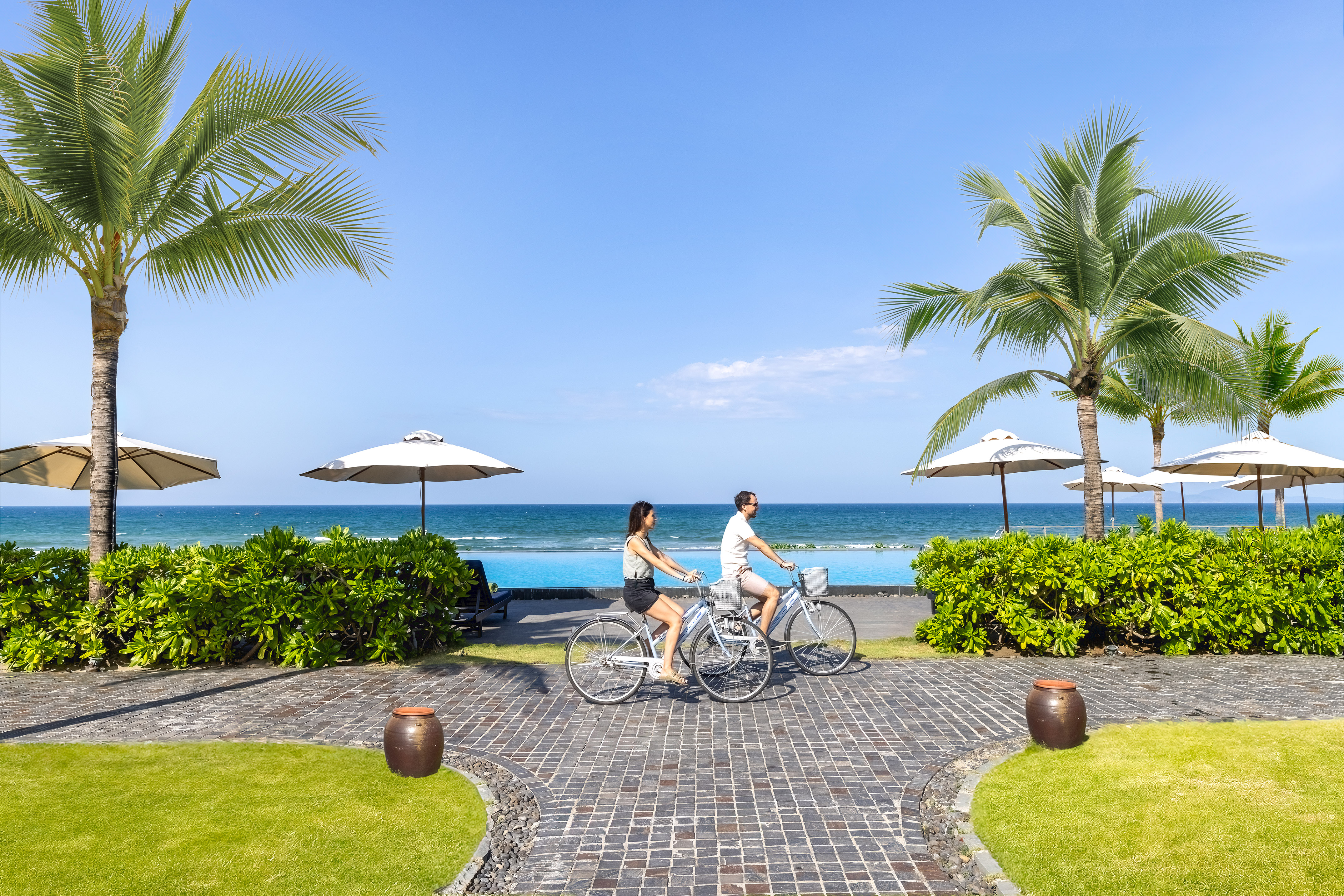 a man and woman riding bicycles on a brick path with palm trees and a body of water