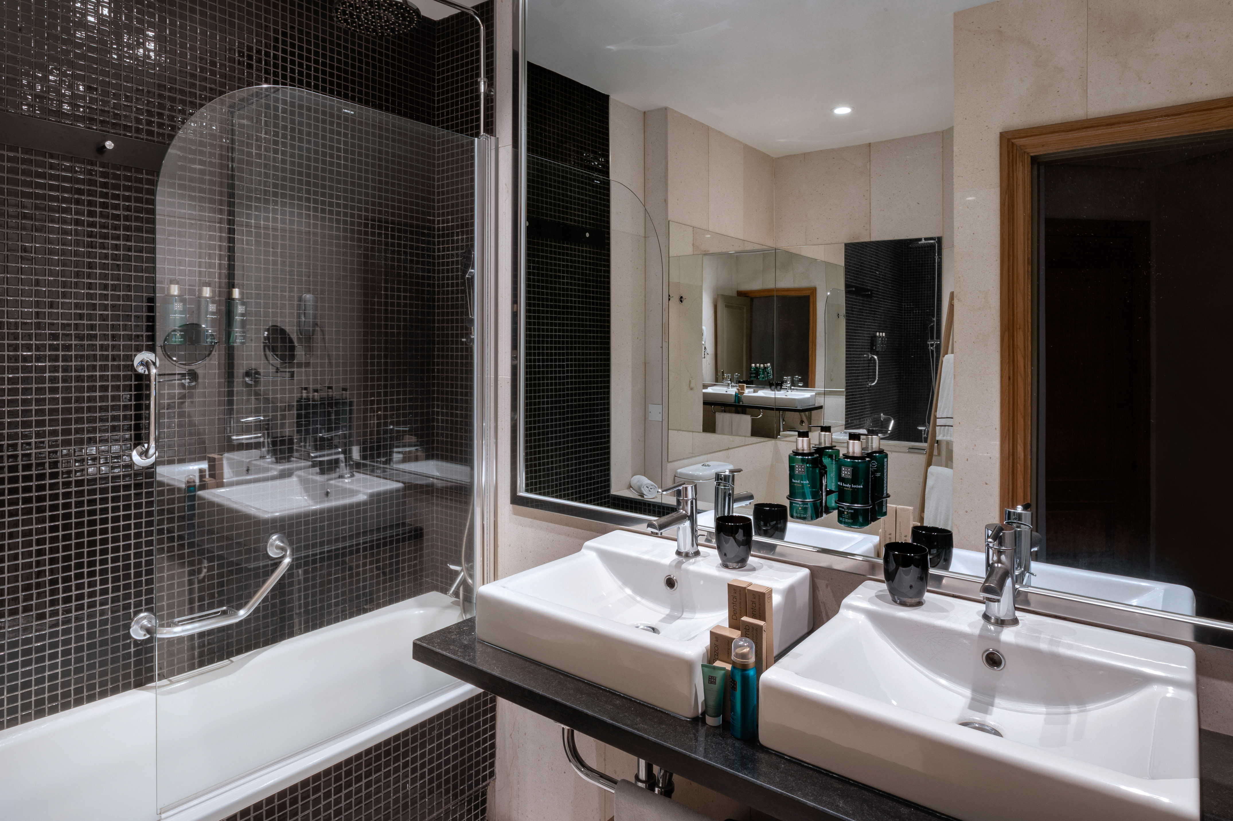 Modern hotel bathroom with black tiled shower-bathtub, glass partition, and double vanity.