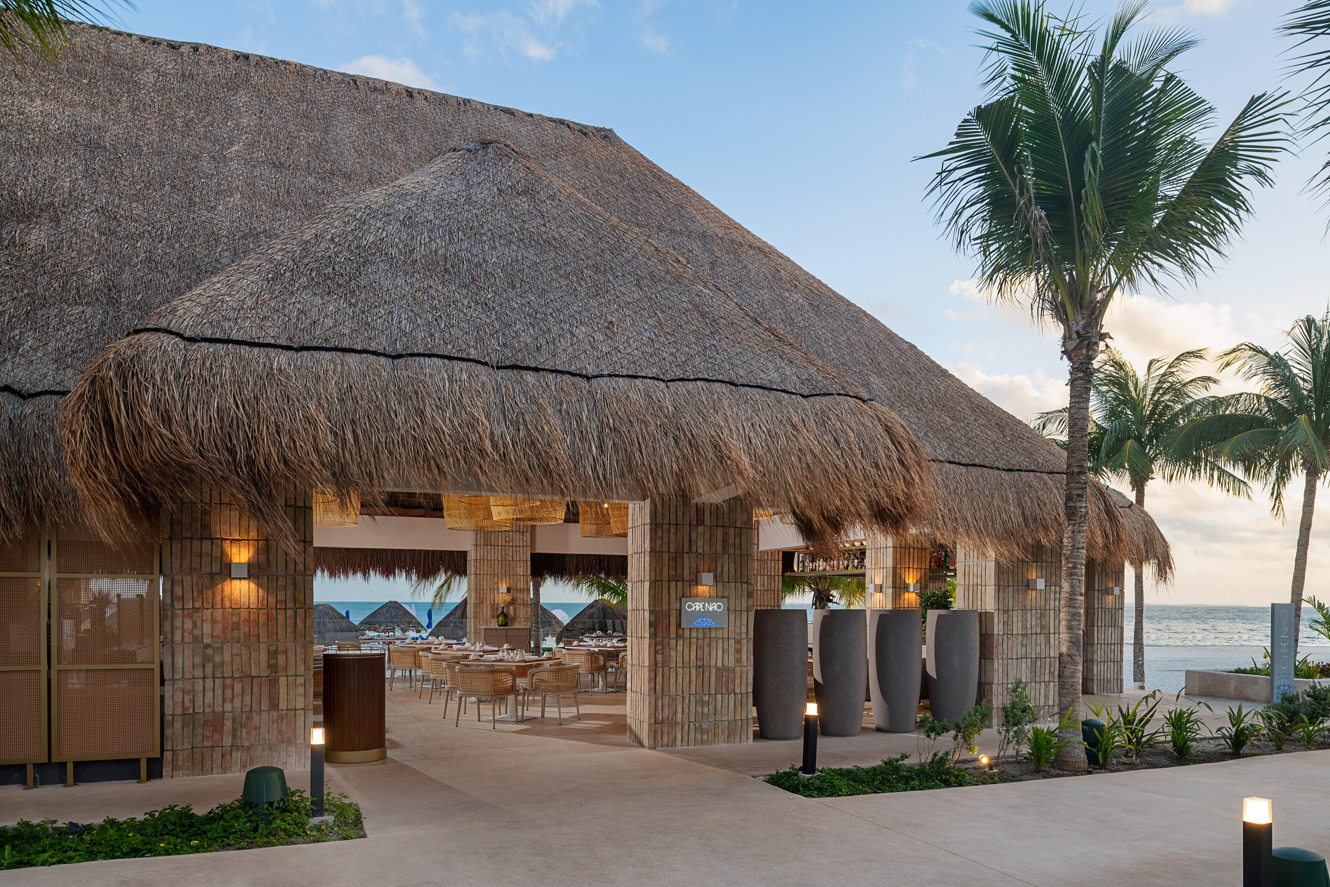 Thatched roof beachfront restaurant with open-air dining, palm trees, and ocean view.