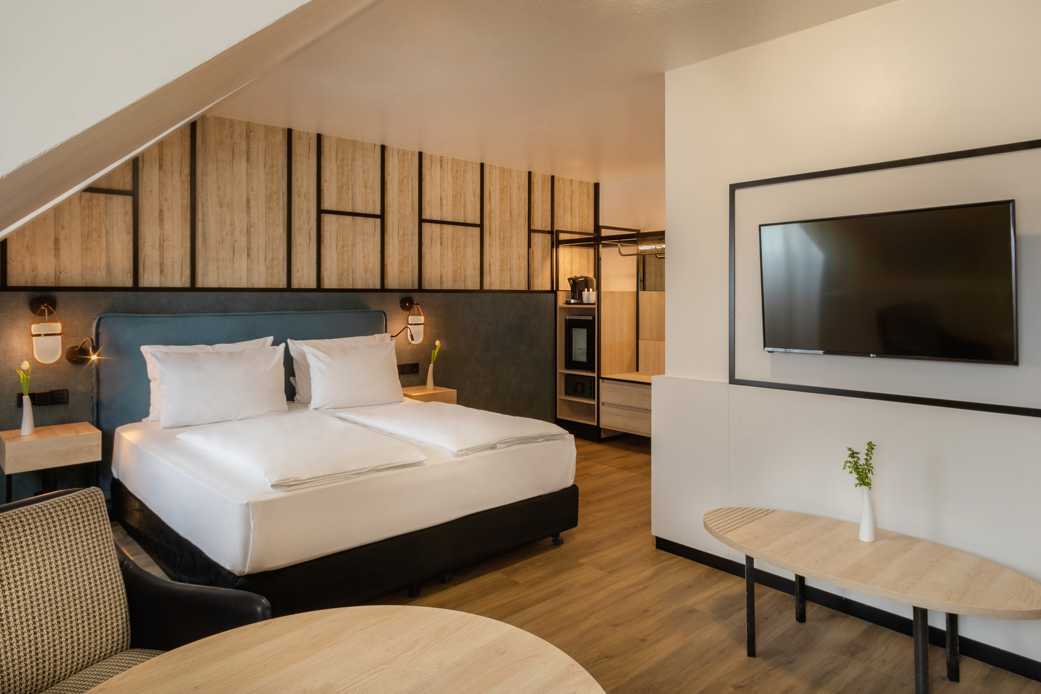 Modern hotel bedroom with queen bed, wood paneling, wall TV, and attic ceiling.