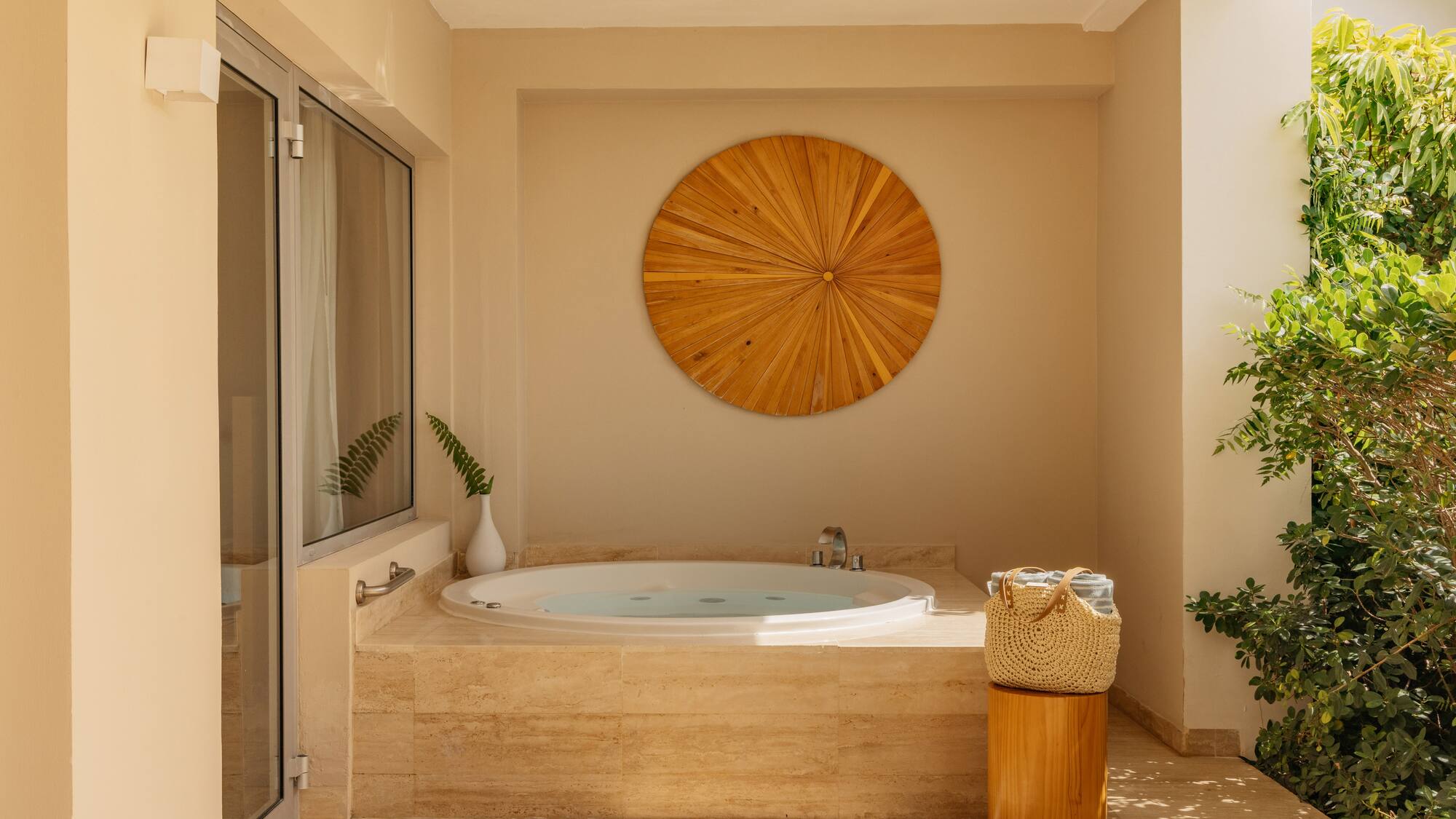 Outdoor hot tub on a sunny patio with wooden wall art and tropical plants.