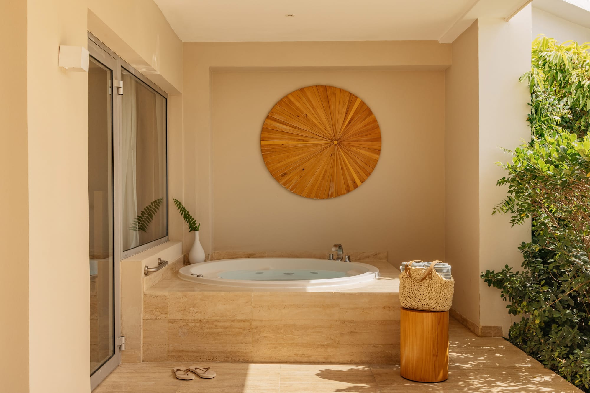 Outdoor hot tub on a sunny patio with wooden wall art and tropical plants.
