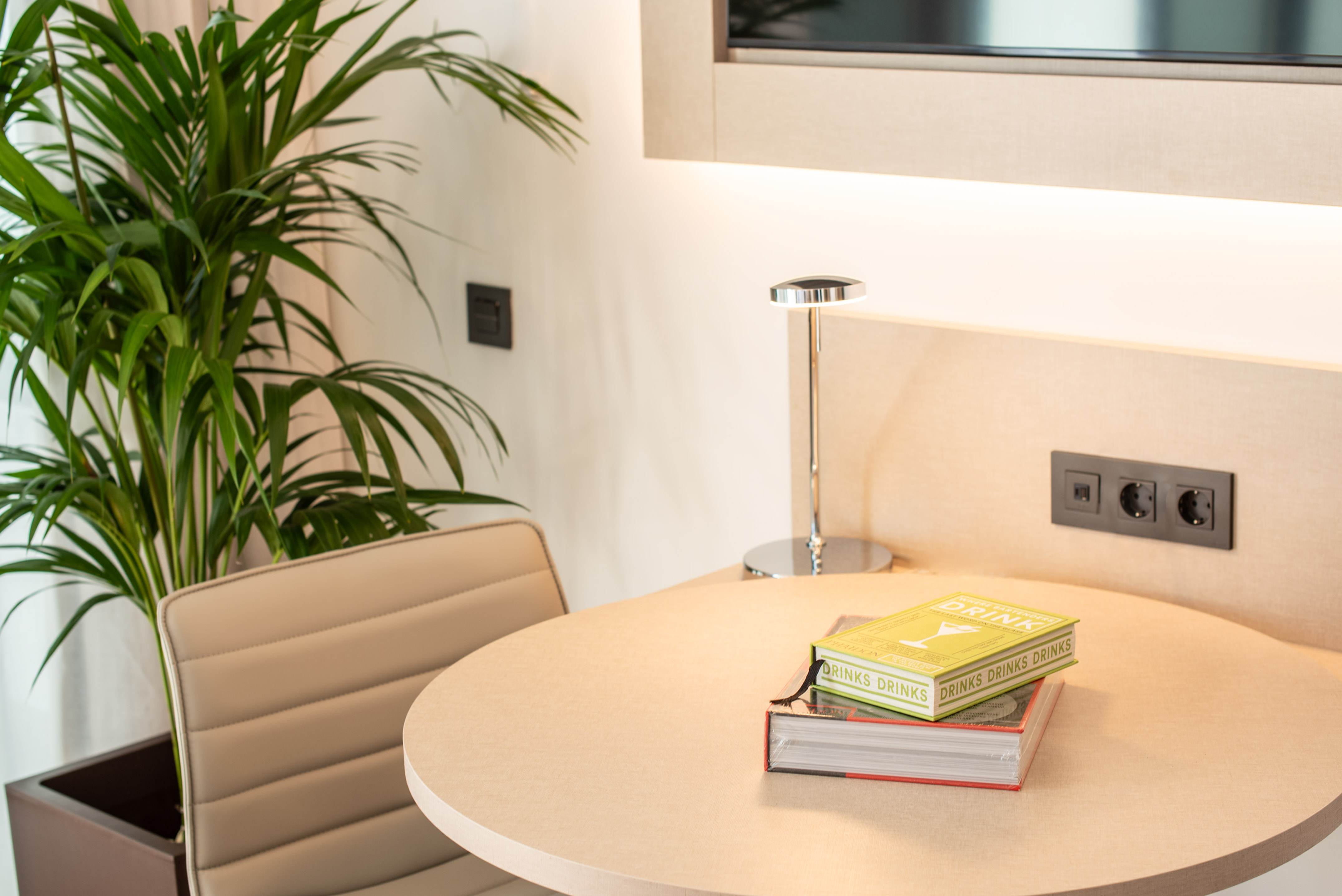 Cozy hotel room desk with books, lamp, beige chair, and large plant.