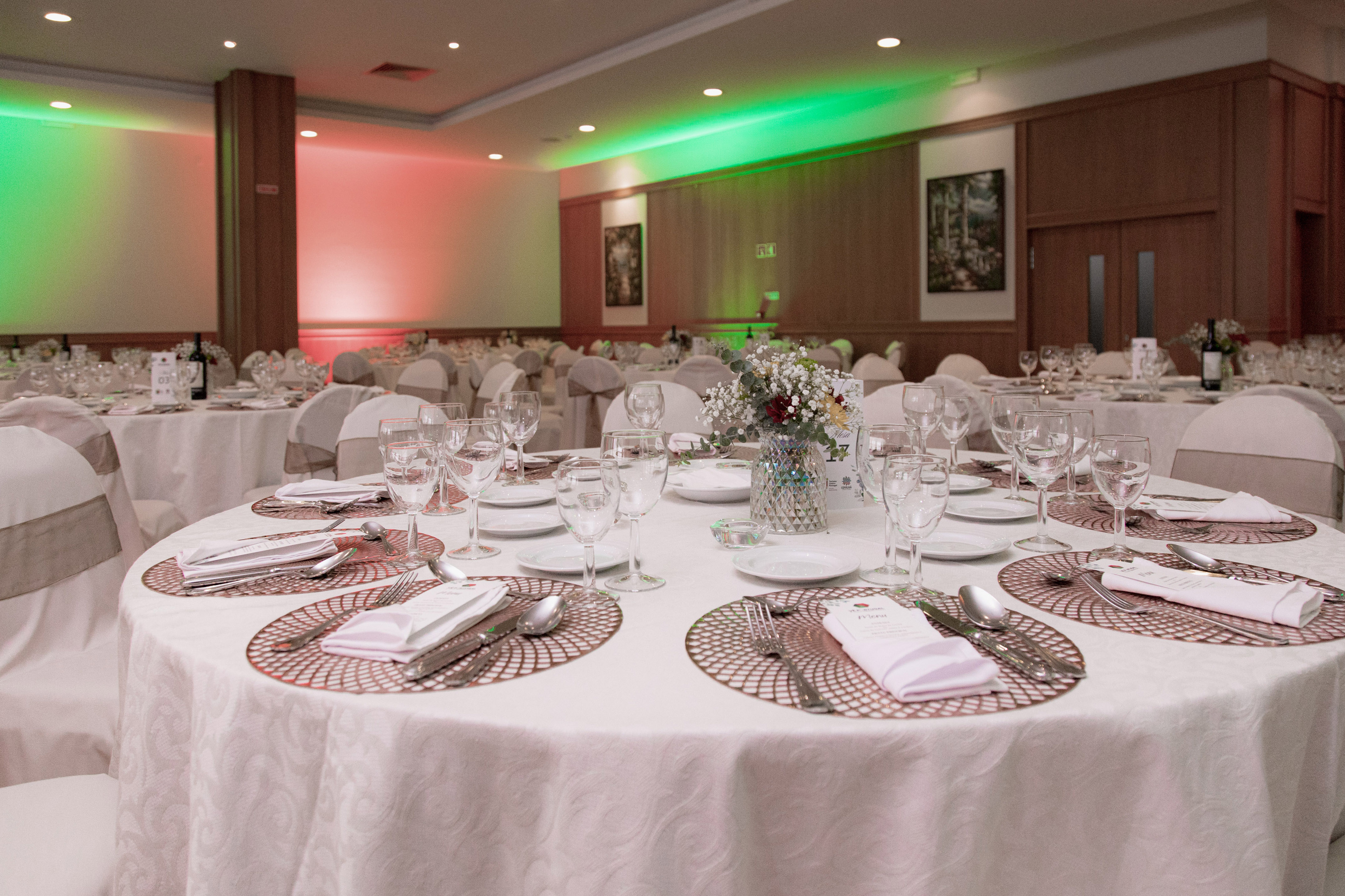 Elegant banquet hall, tables set with flowers, glasses, and red-green lighting.
