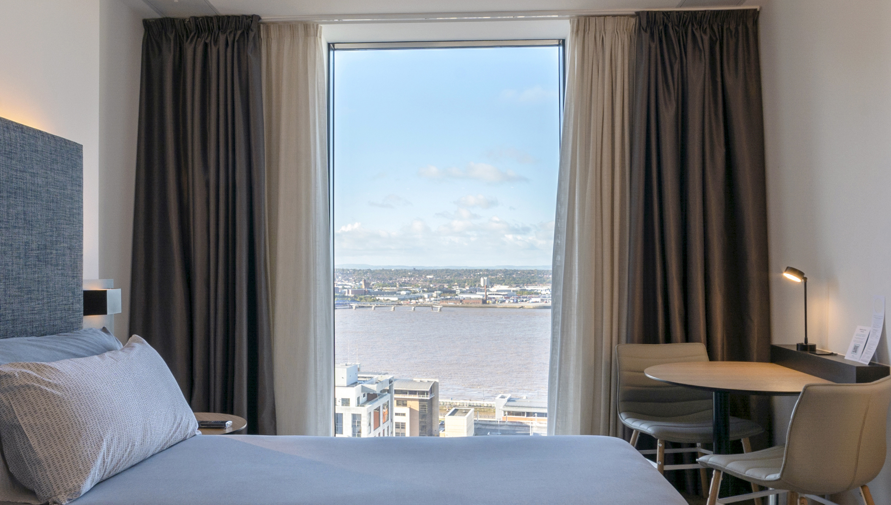 Hotel room with bed, table, and panoramic window view of a river and city.