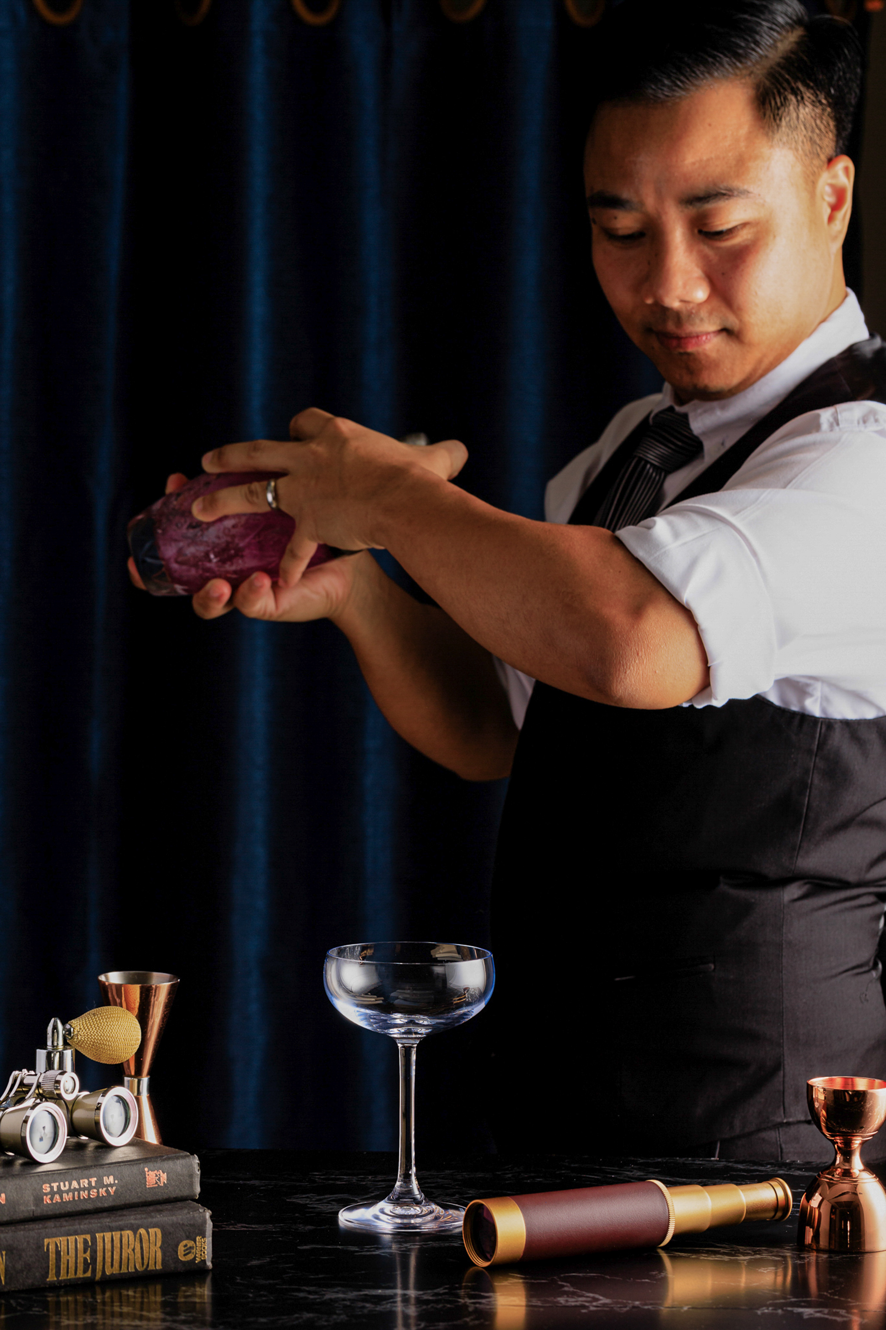 Bartender preparing vibrant purple cocktail, surrounded by vintage bar items.