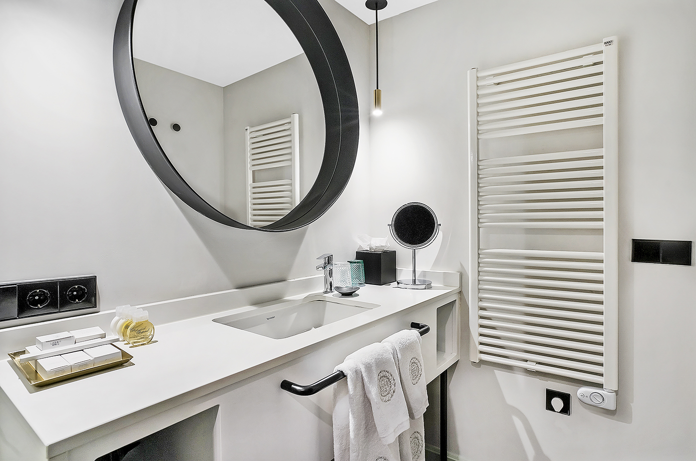 Contemporary hotel bathroom with white vanity, round mirror, and towel radiator.
