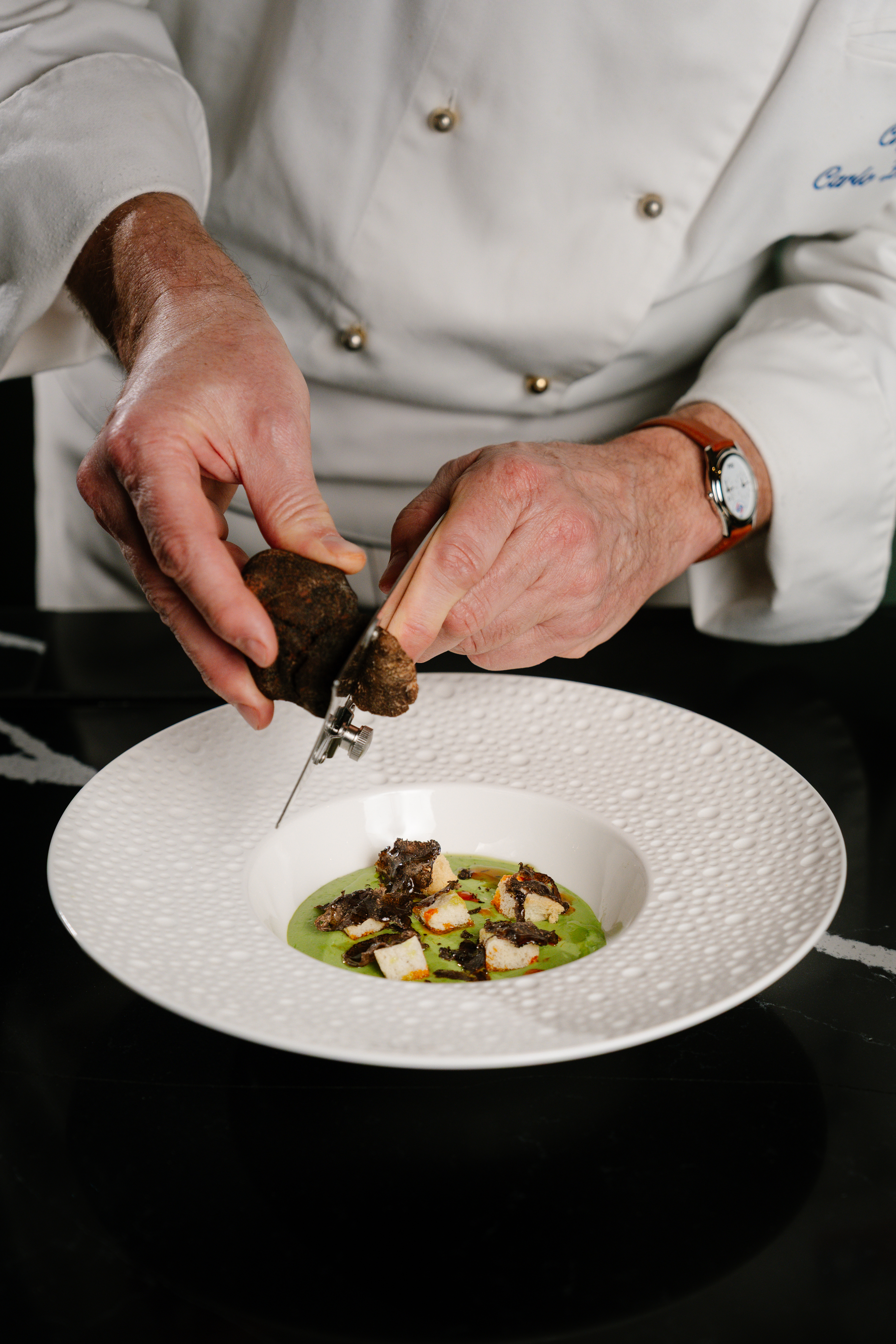 Chef shaving black truffle onto a gourmet dish with green puree and croutons.