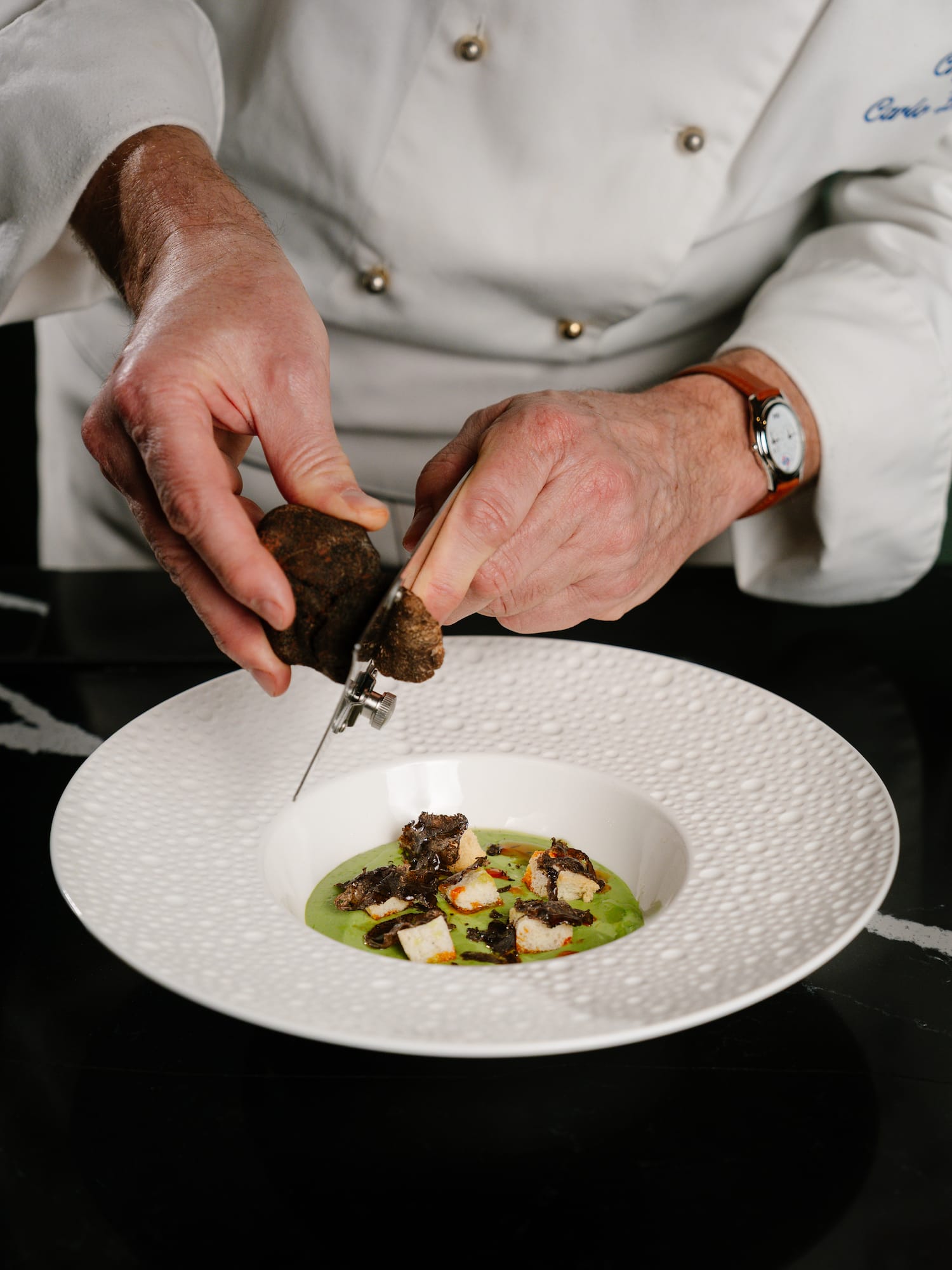 Chef shaving black truffle onto a gourmet dish with green puree and croutons.