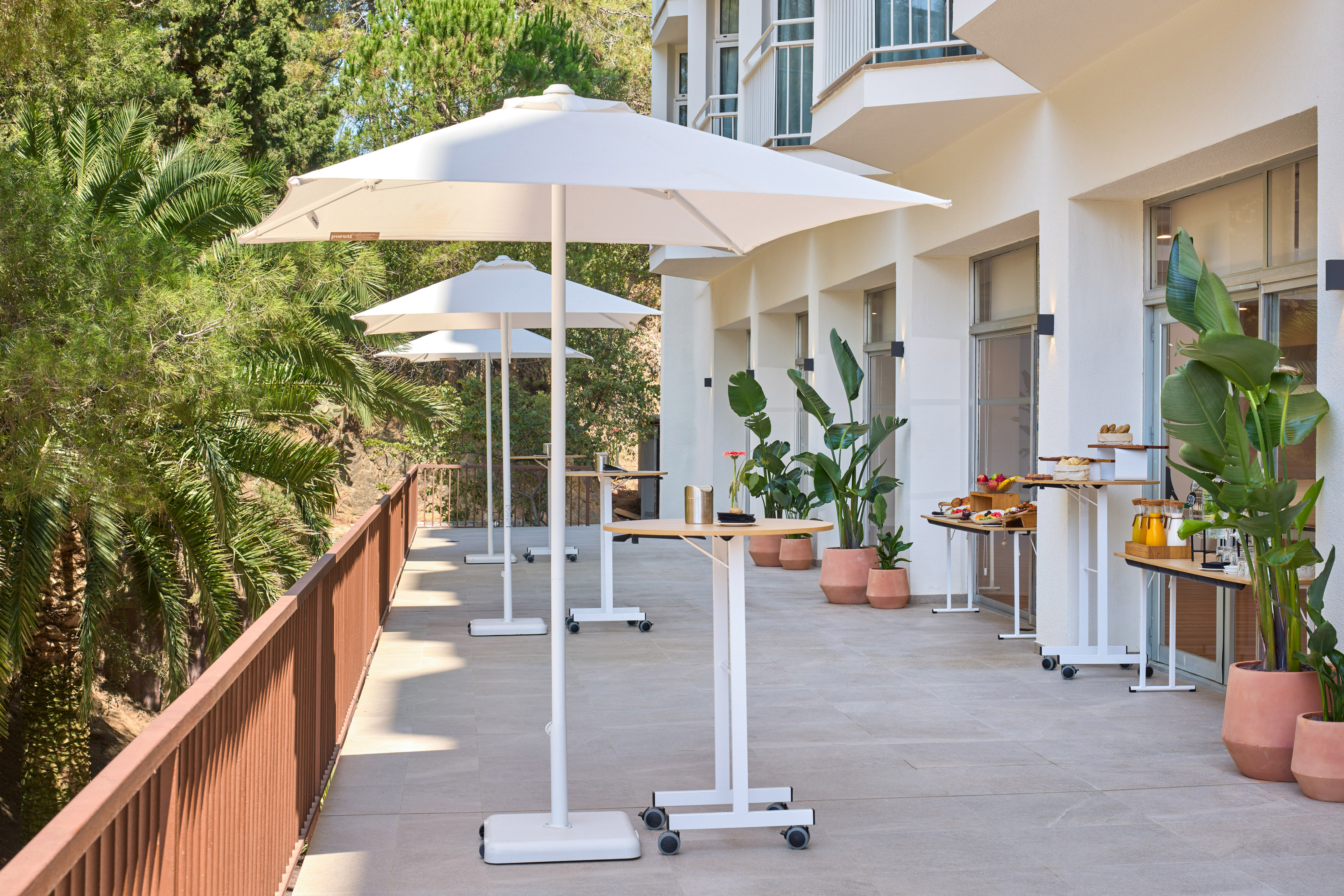 Sunny outdoor patio with white umbrellas, buffet tables, and lush green plants.