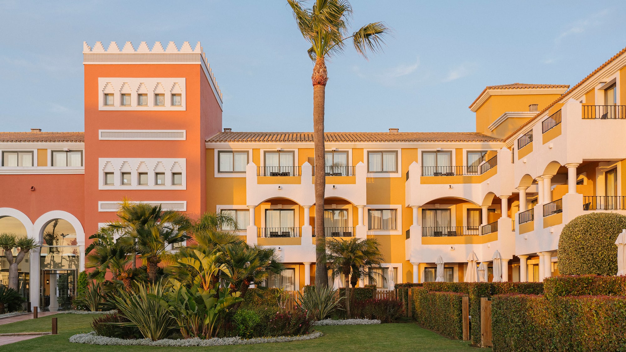 Vibrant orange/yellow Mediterranean hotel with palm trees and green lawn.