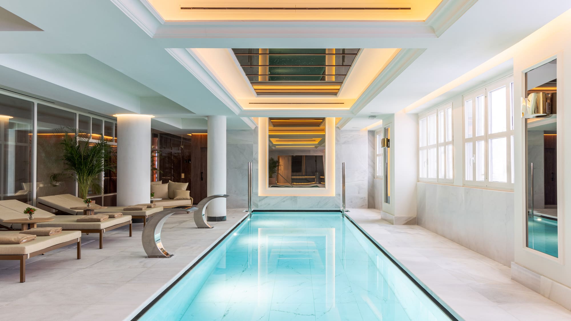 Serene indoor hotel pool with marble surrounds, loungers, and warm lighting.