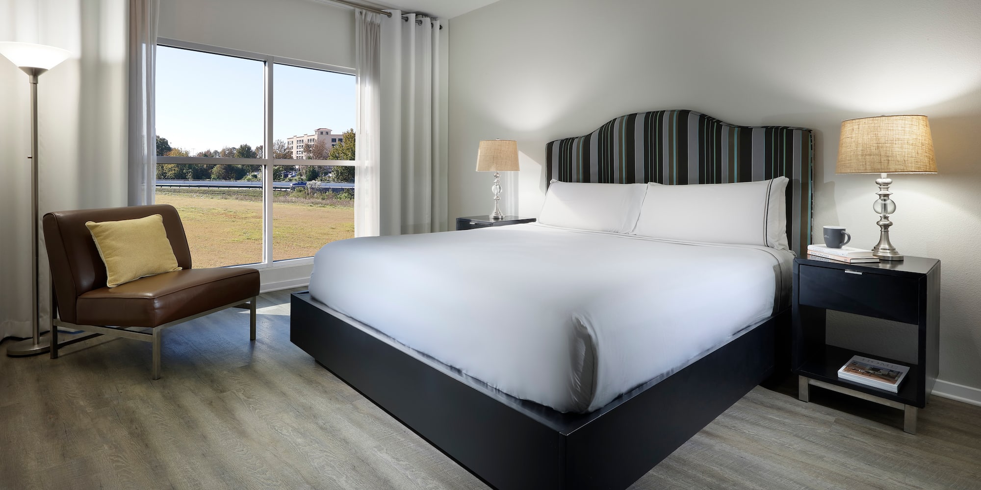 Modern hotel bedroom with king bed, striped headboard, wood floor, and window view.