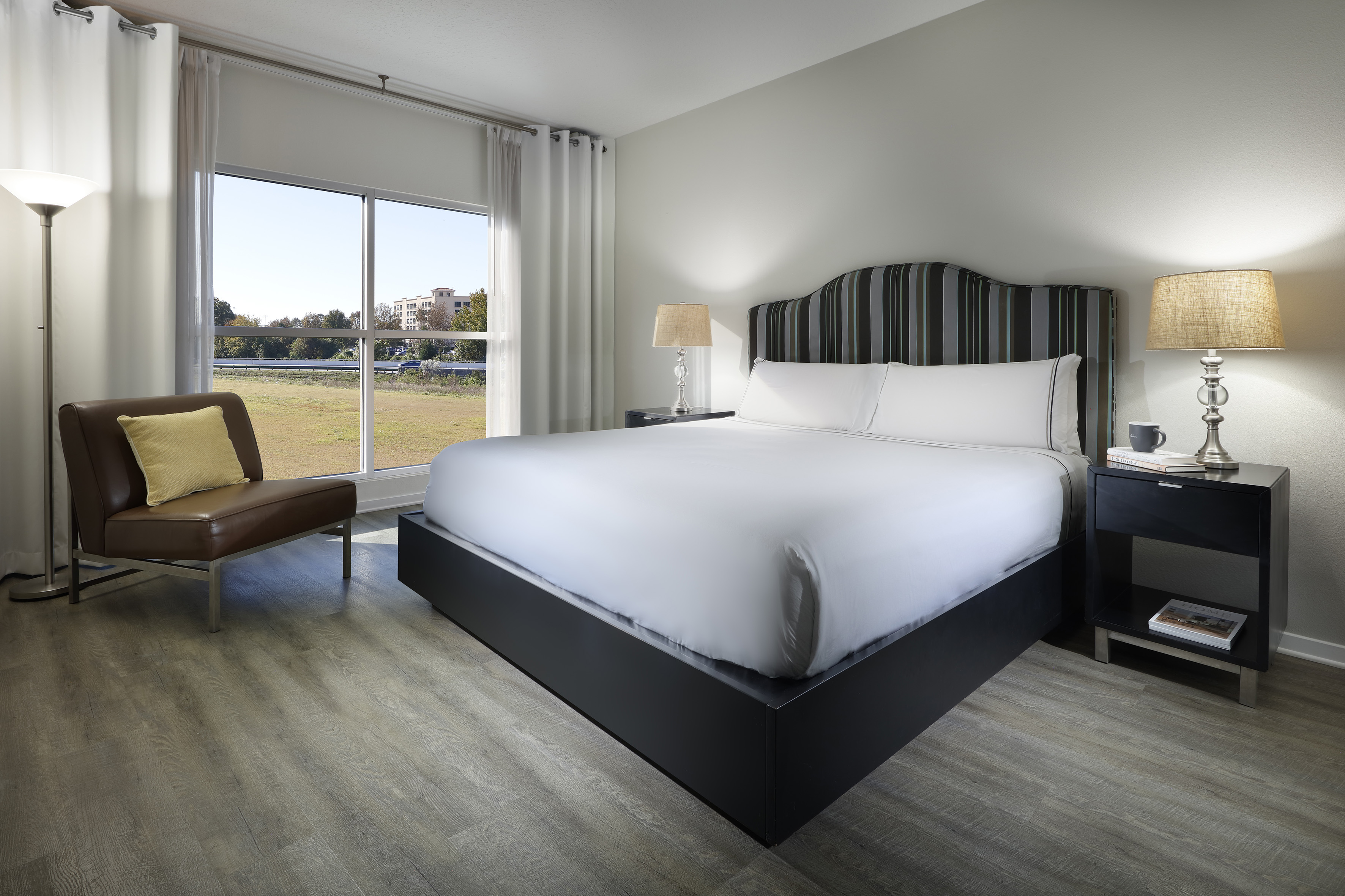 Modern hotel bedroom with king bed, striped headboard, wood floor, and window view.