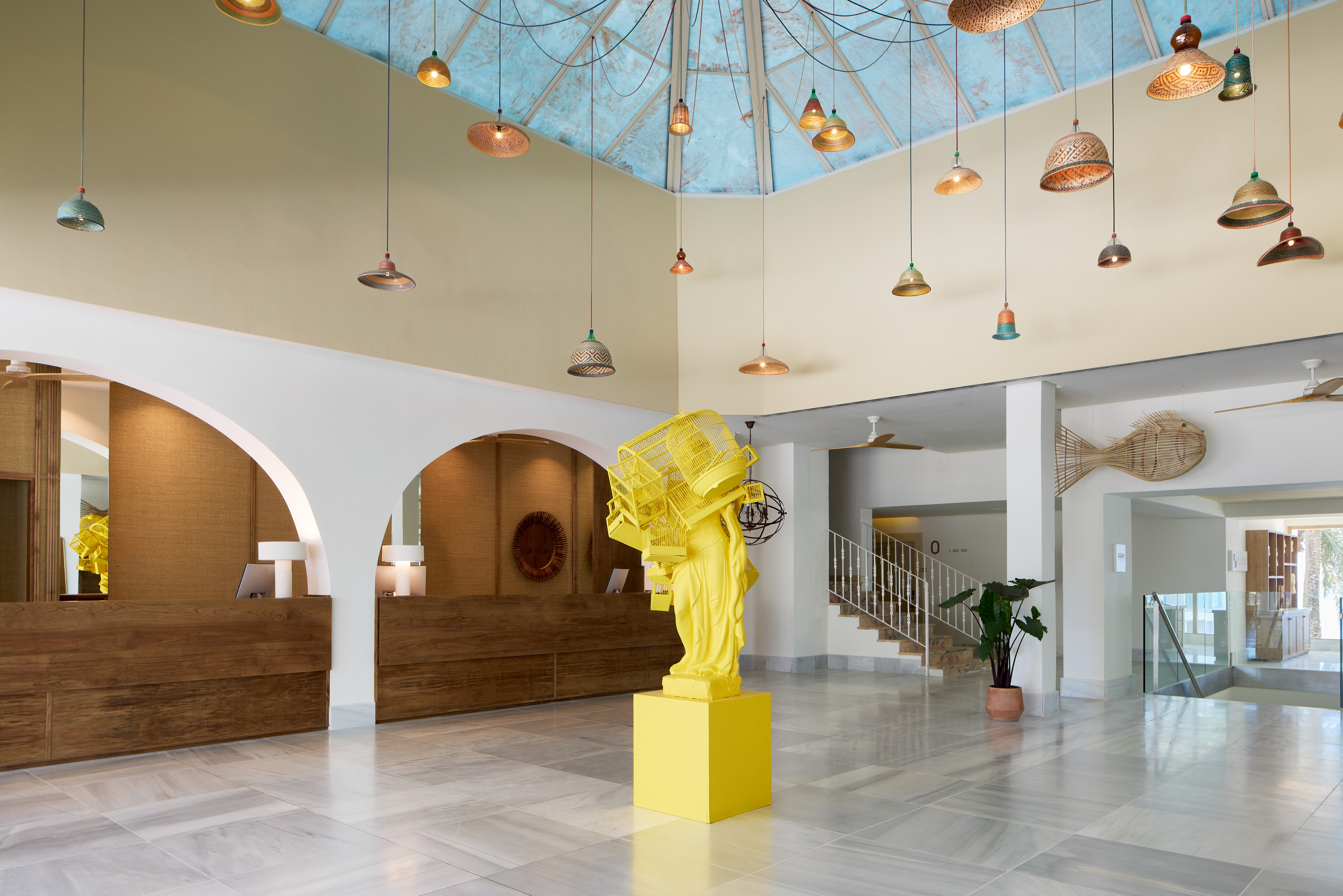 Modern hotel lobby with yellow sculpture, wooden reception, colorful hanging lights.