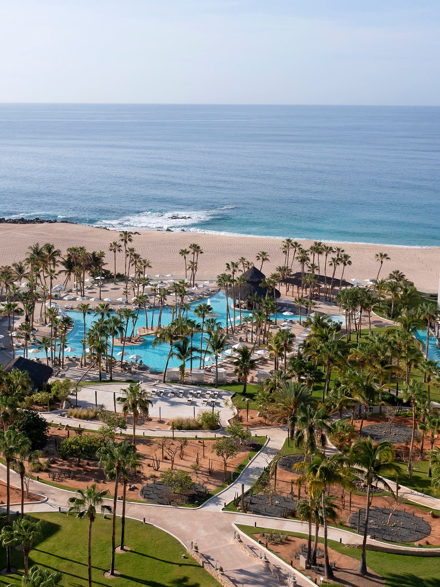 Aerial view of luxury beachfront resort with large swimming pool, palm trees, and ocean.