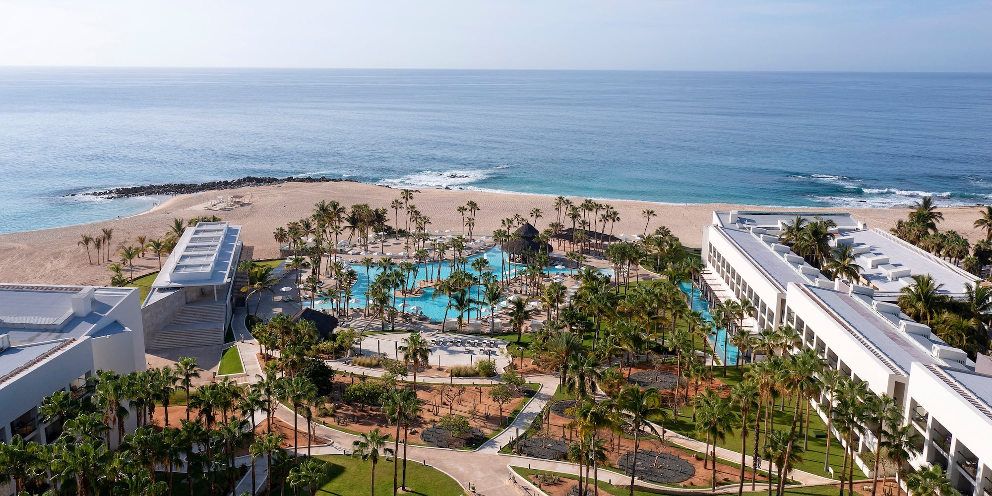 Aerial view of luxury beachfront resort with large swimming pool, palm trees, and ocean.