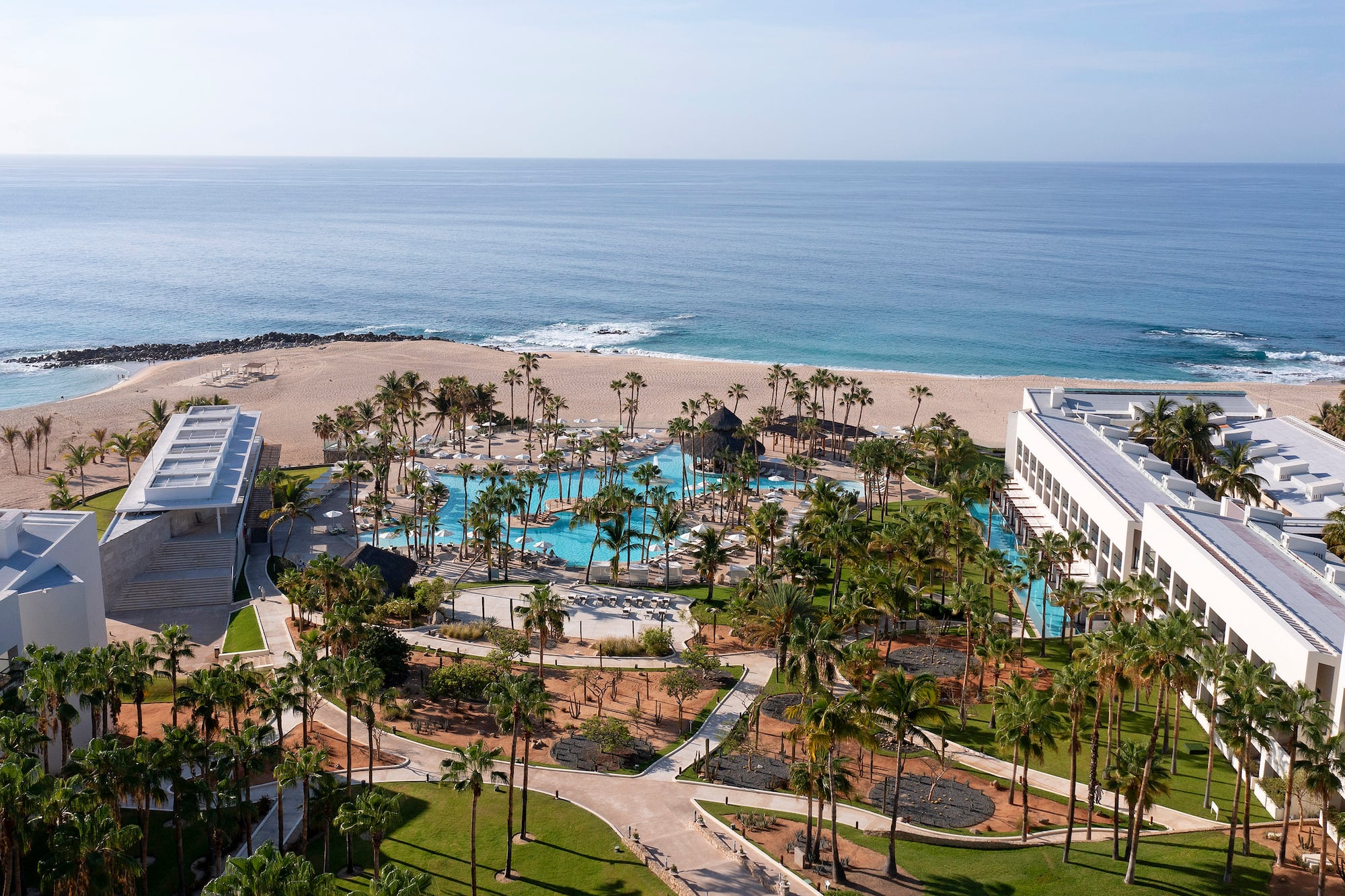 Aerial view of luxury beachfront resort with large swimming pool, palm trees, and ocean.