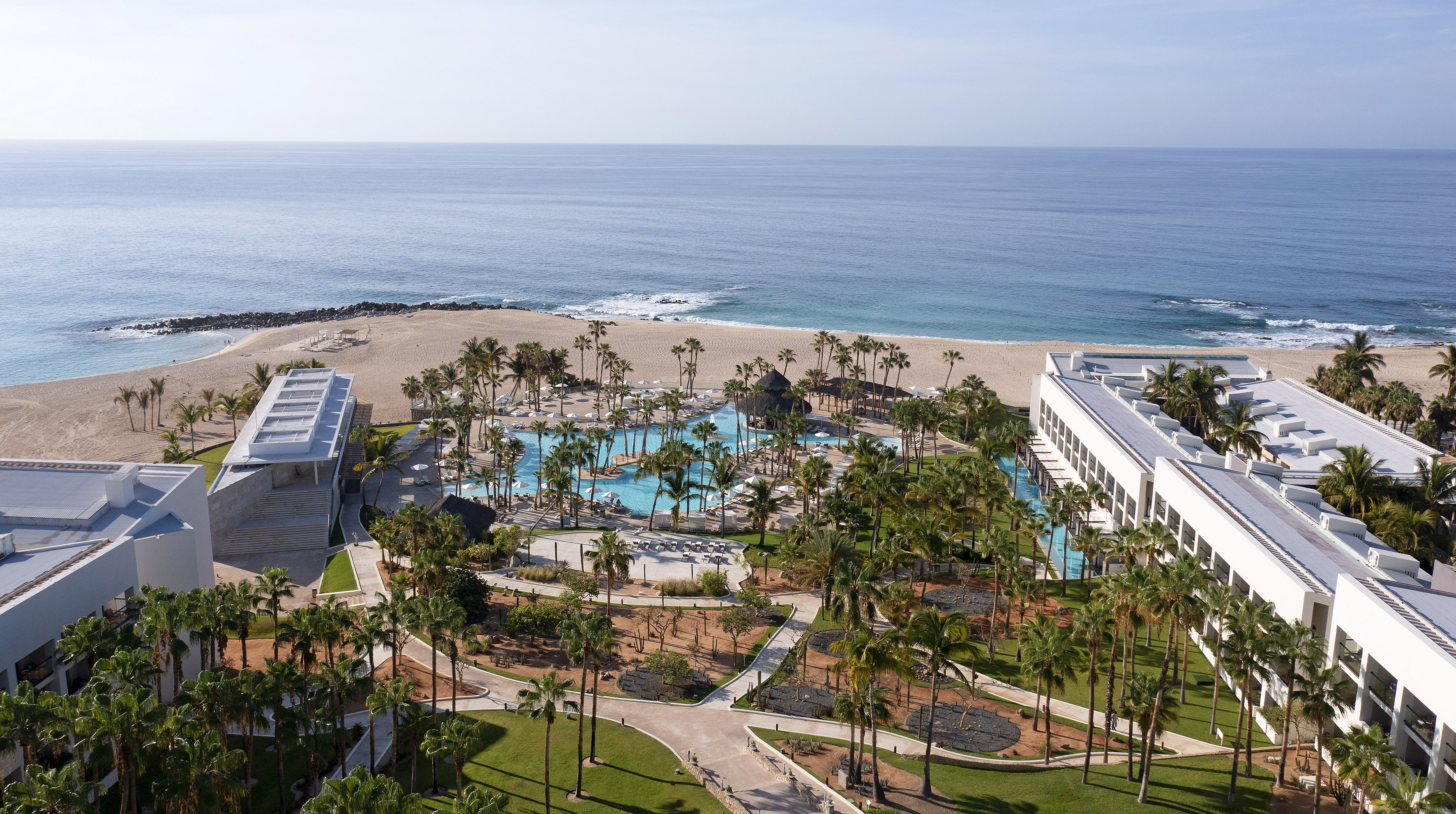 Aerial view of luxury beachfront resort with large swimming pool, palm trees, and ocean.