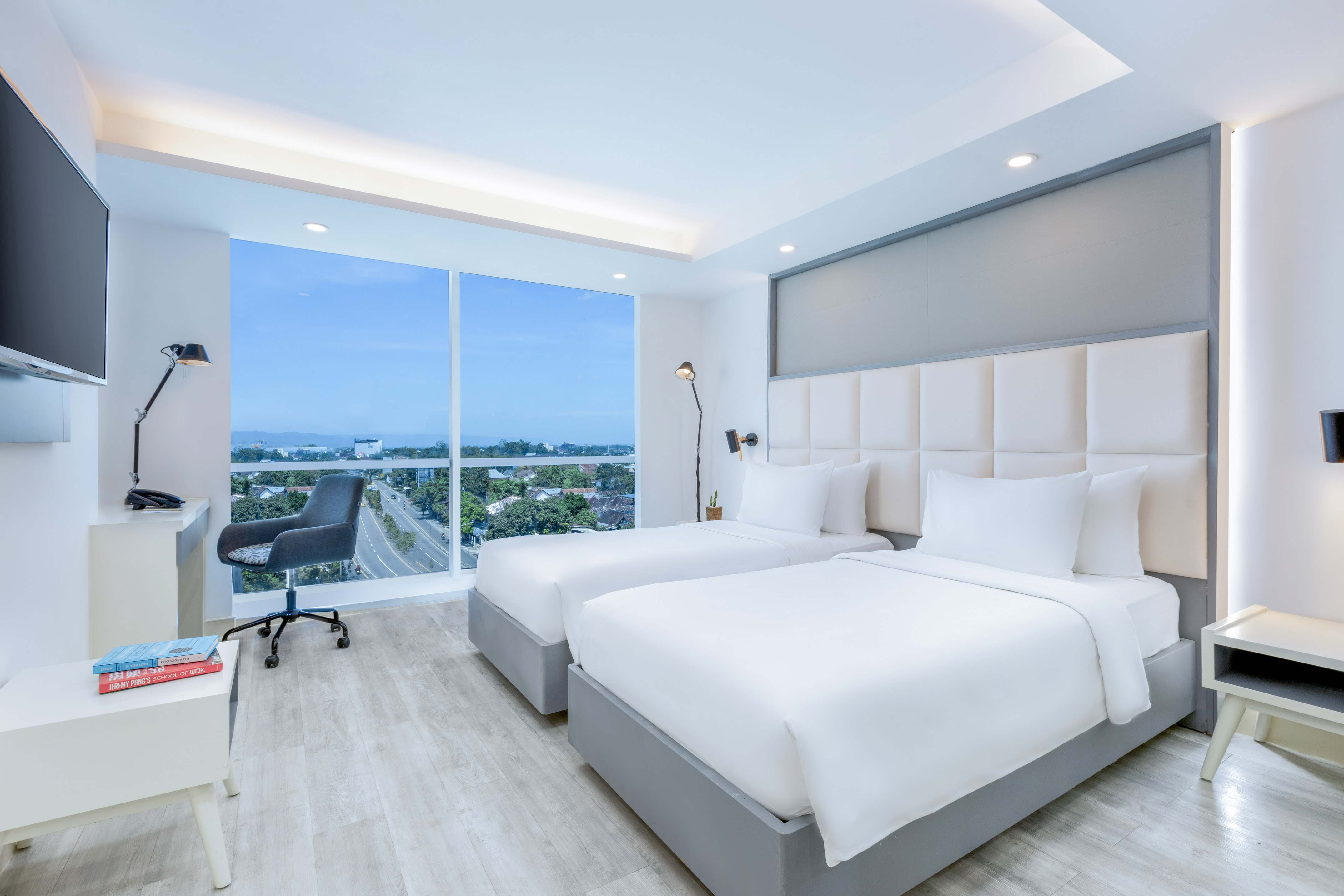 Bright modern hotel room with two beds, desk, and large window city view.