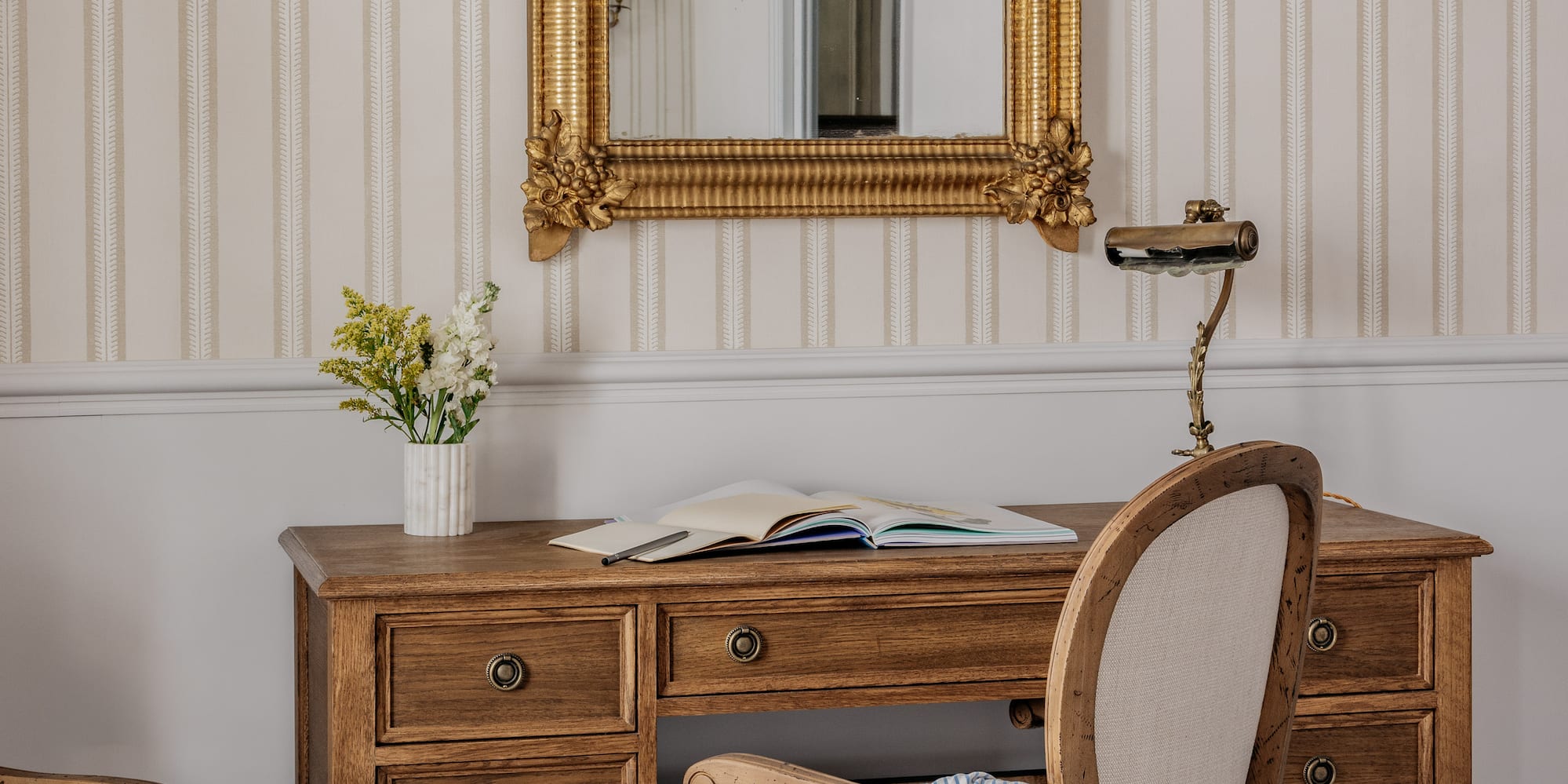 Vintage wooden desk, ornate gold mirror, classic chair, striped wallpaper, checkered floor.