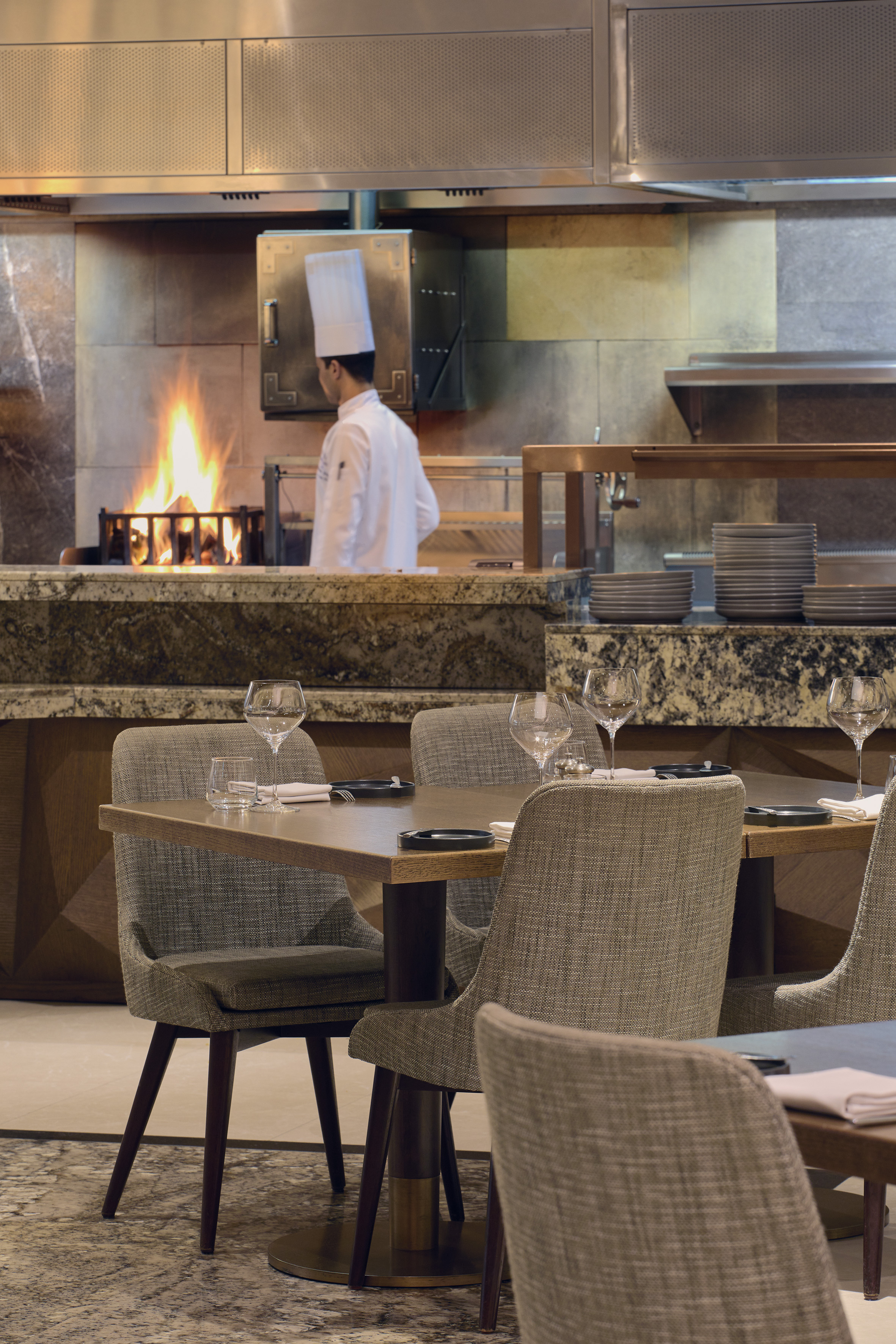 Open kitchen restaurant with chef at wood-fired grill, set dining tables.