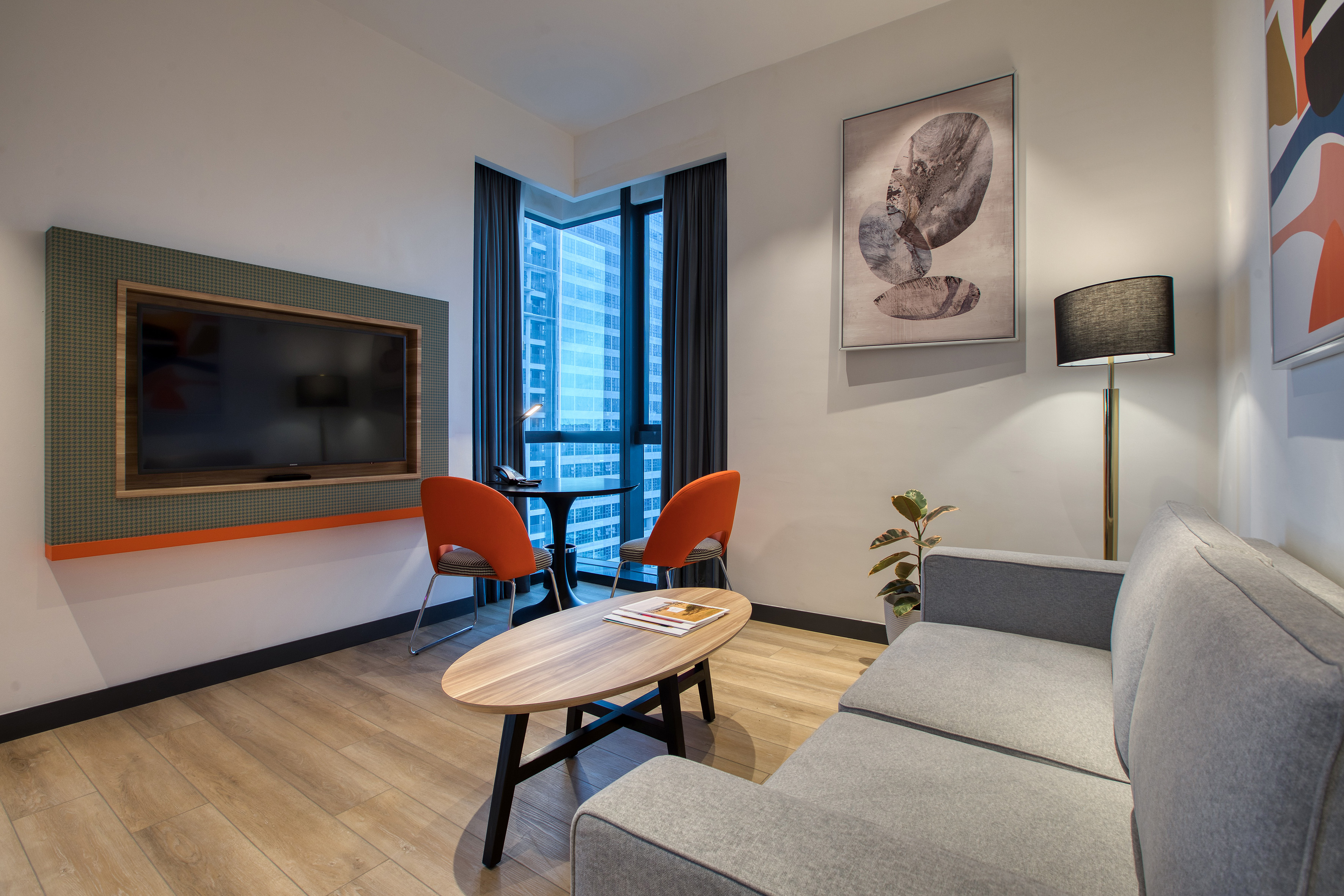 Contemporary hotel room with gray sofa, TV, orange chairs, city view.