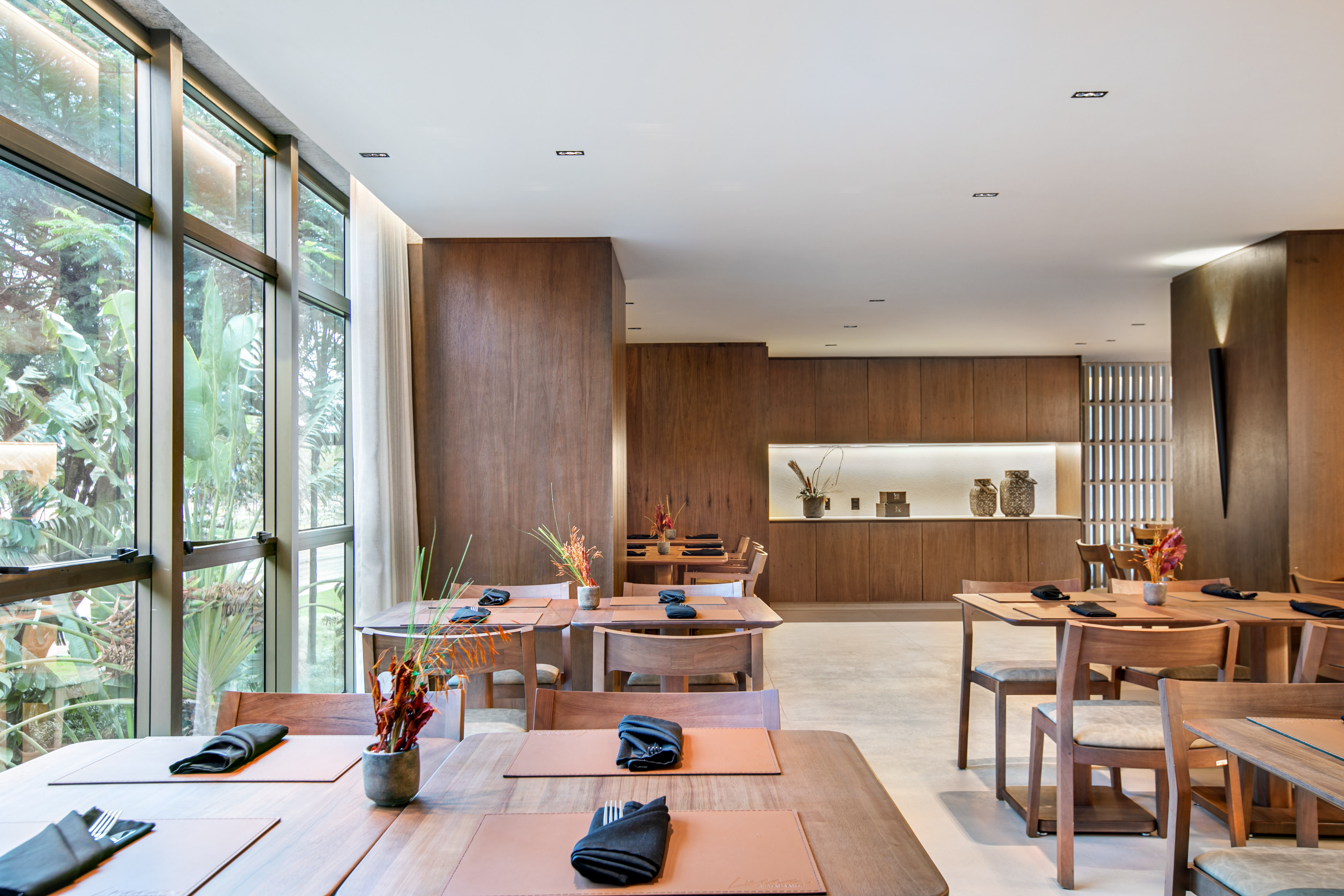 Modern restaurant with wooden tables, large windows, and a lush tropical view.