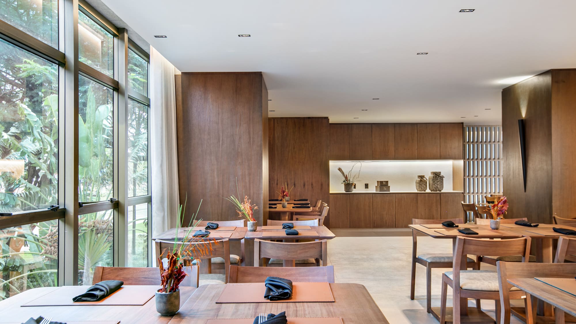 Modern restaurant with wooden tables, large windows, and a lush tropical view.