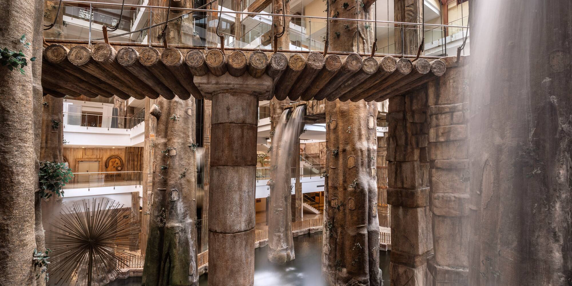 Modern hotel atrium with indoor waterfalls, tree-shaped columns, and log bridge.