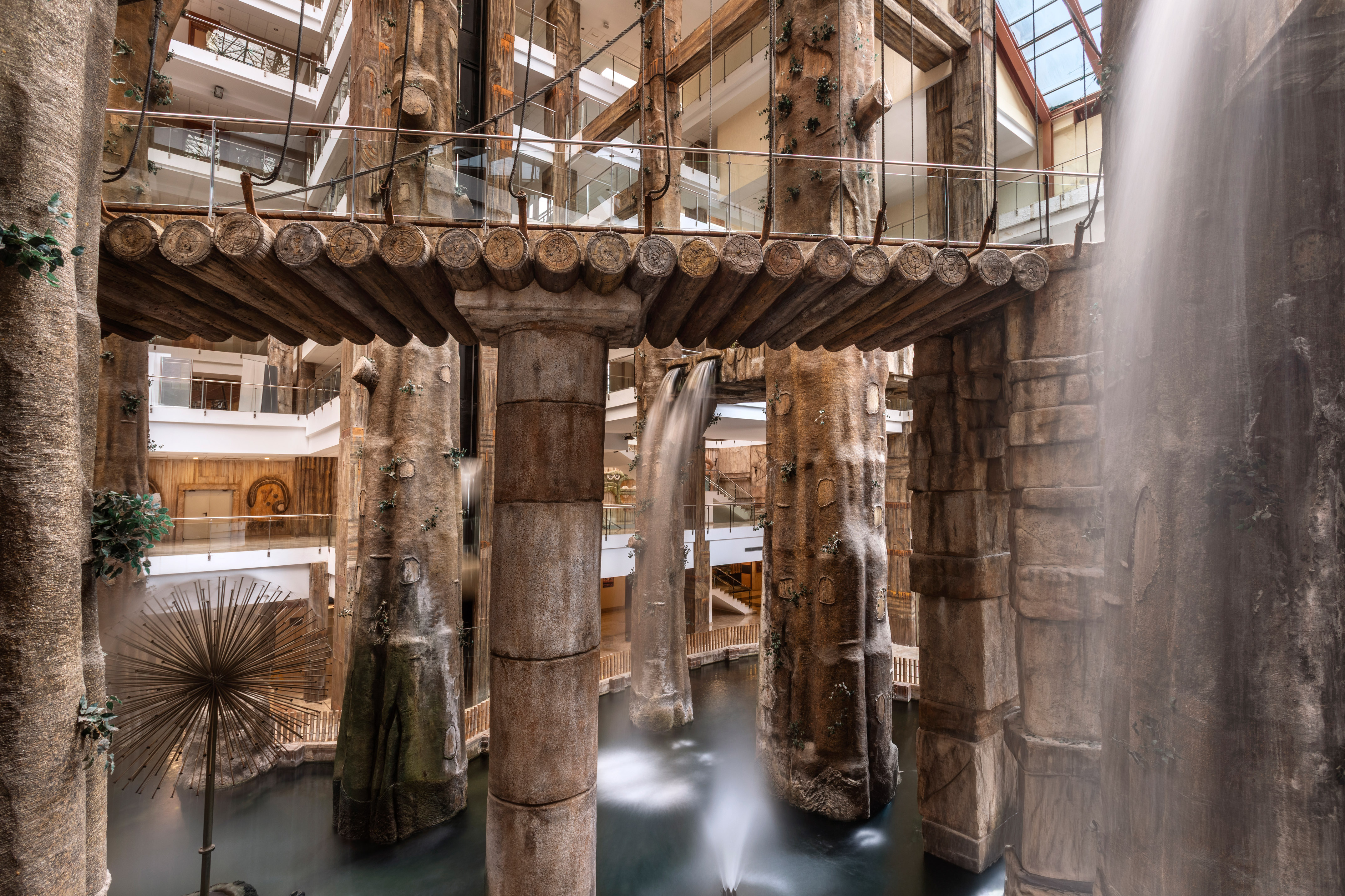 Modern hotel atrium with indoor waterfalls, tree-shaped columns, and log bridge.