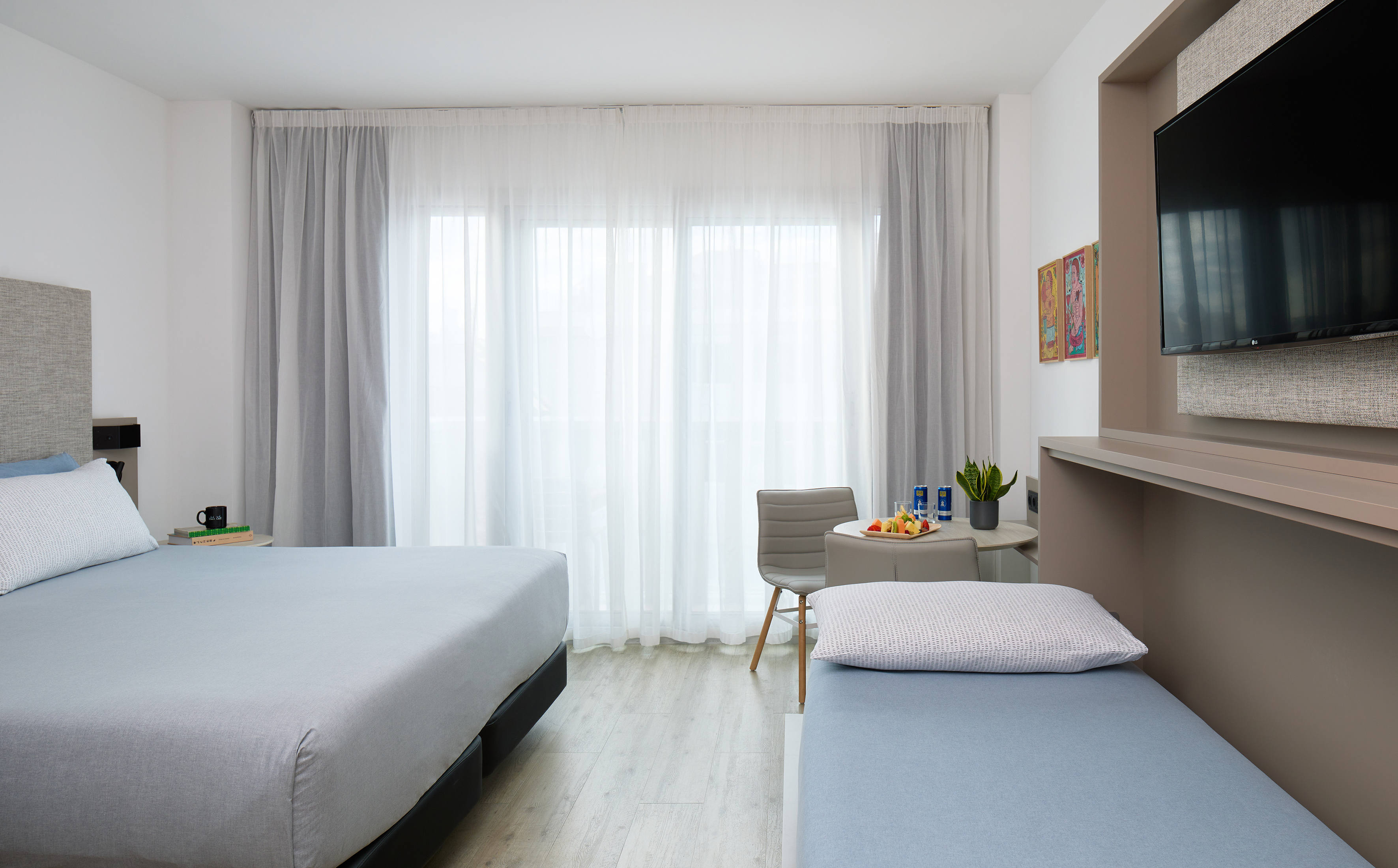 Modern hotel room with two beds, dining table, TV, and large window.