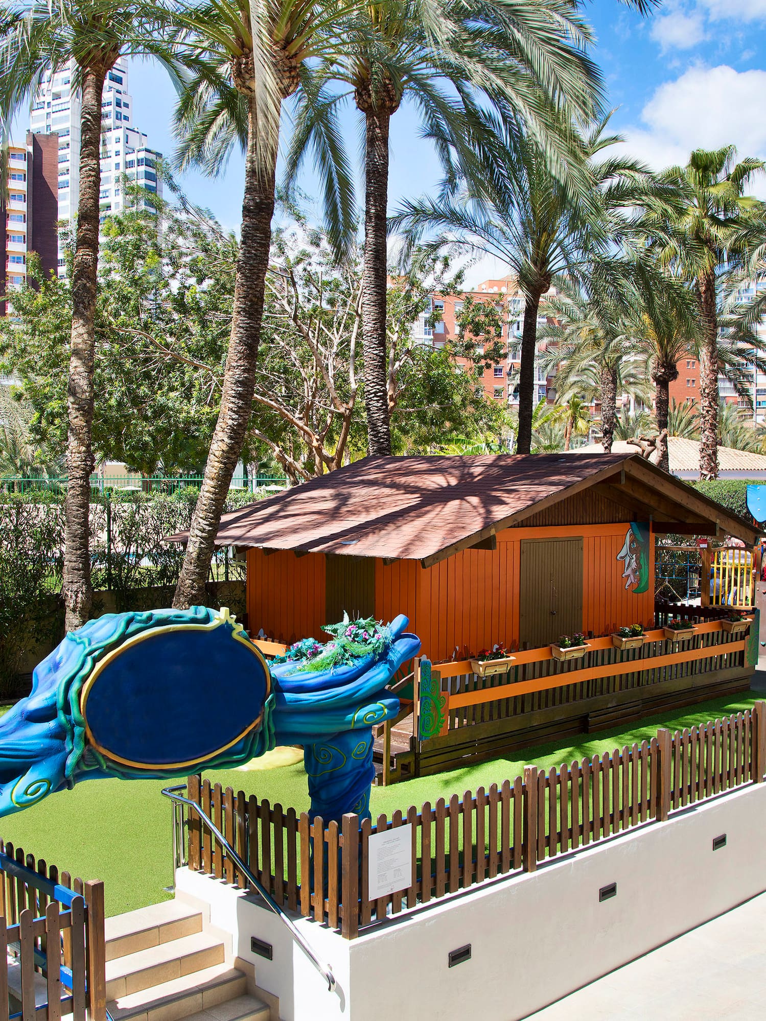 Parque infantil exterior con tobogán, cabaña, césped y palmeras de hotel.