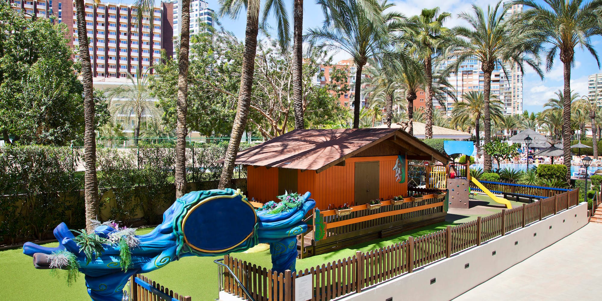 Parque infantil exterior con casita, tobogán, césped y palmeras en hotel.