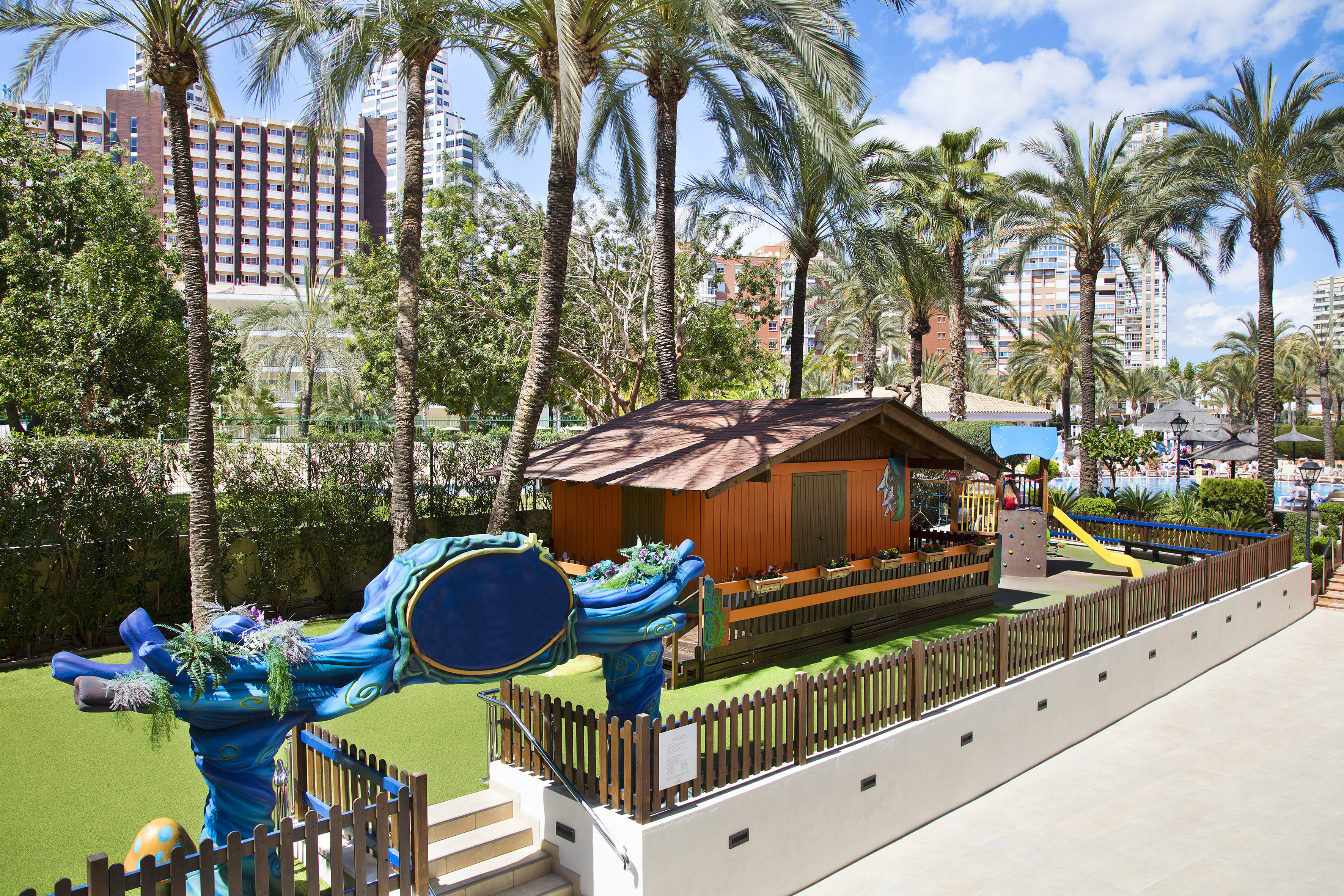 Parque infantil exterior con tobogán, cabaña, césped y palmeras de hotel.