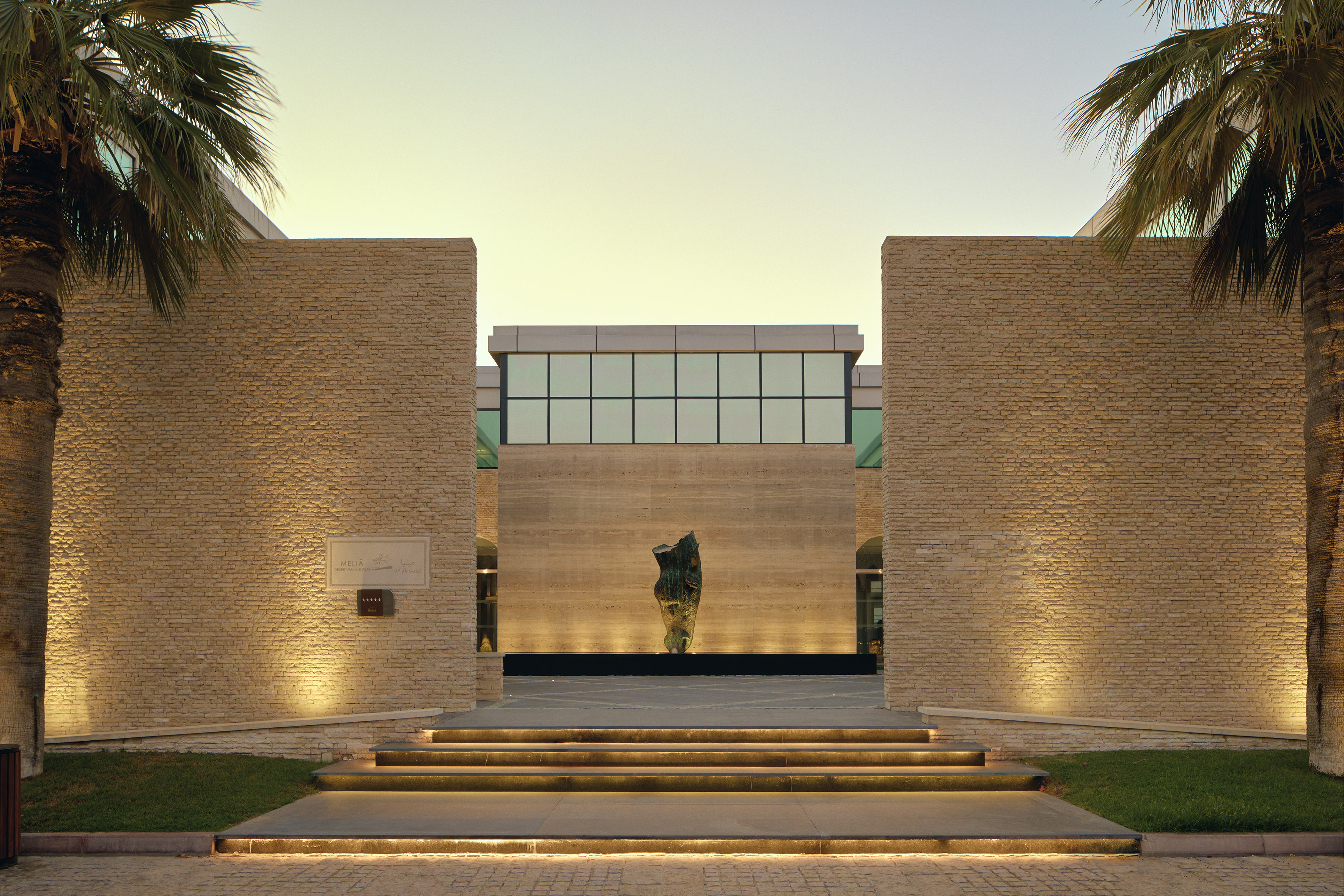 Meliá hotel entrance, stone facade, palm trees, lit steps, modern sculpture.