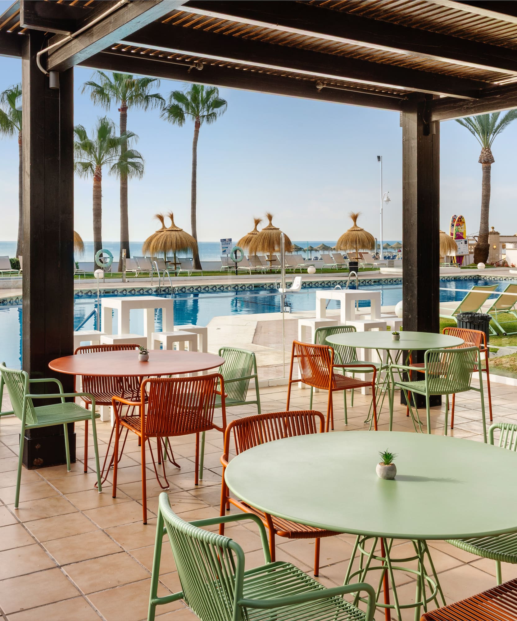 Terraza cubierta de hotel con piscina, playa, palmeras y mobiliario moderno.