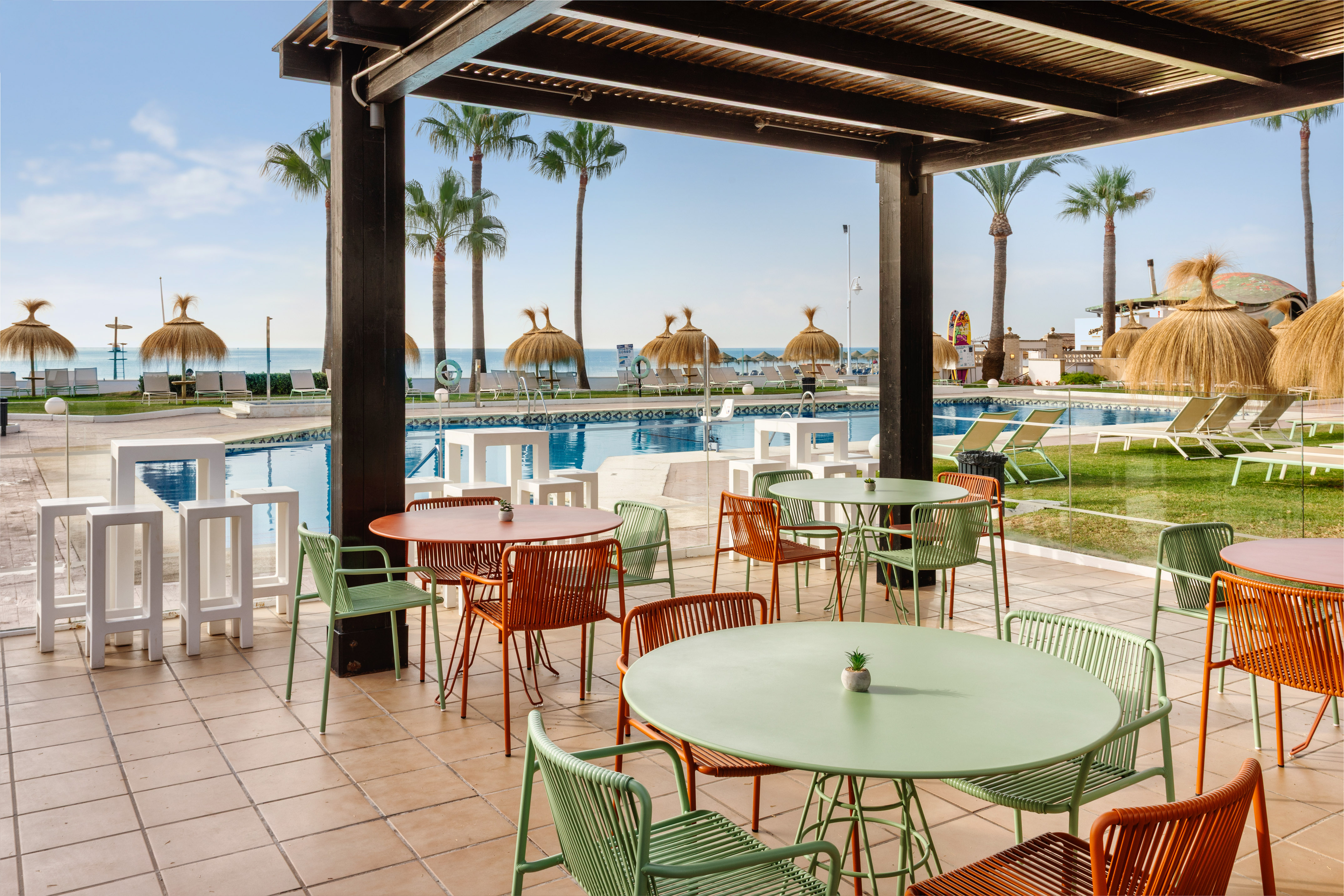Terraza cubierta de hotel con piscina, playa, palmeras y mobiliario moderno.