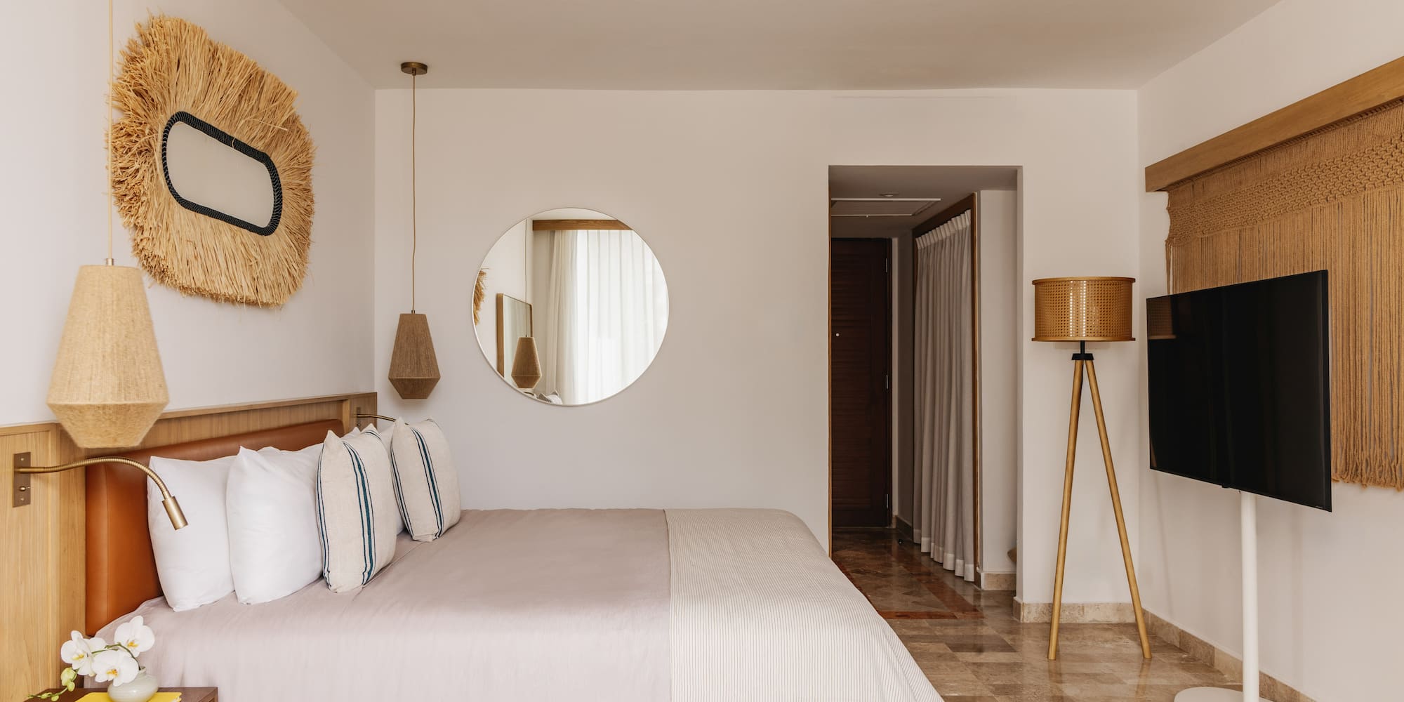 Modern hotel bedroom with large bed, natural decor, round mirror, and TV.