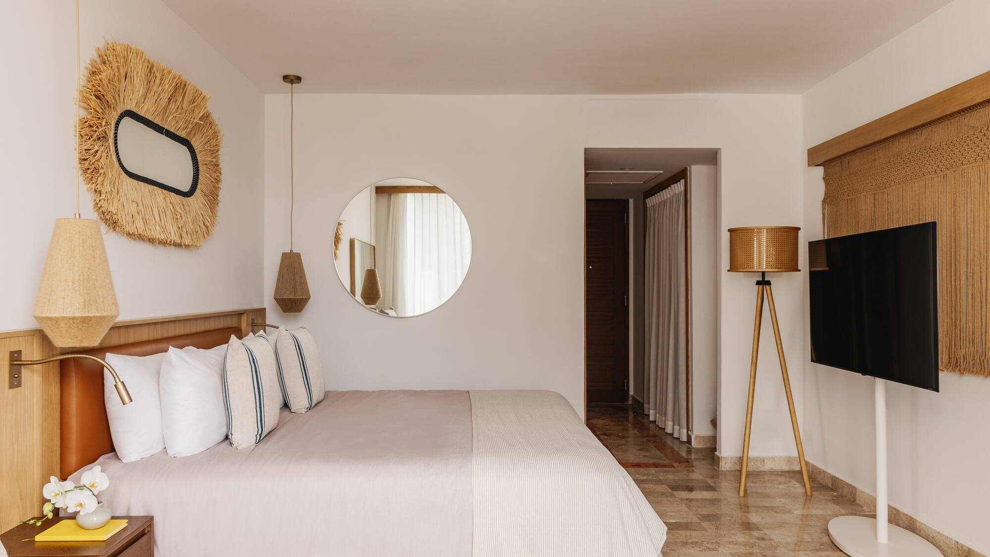 Modern hotel bedroom with large bed, natural decor, round mirror, and TV.