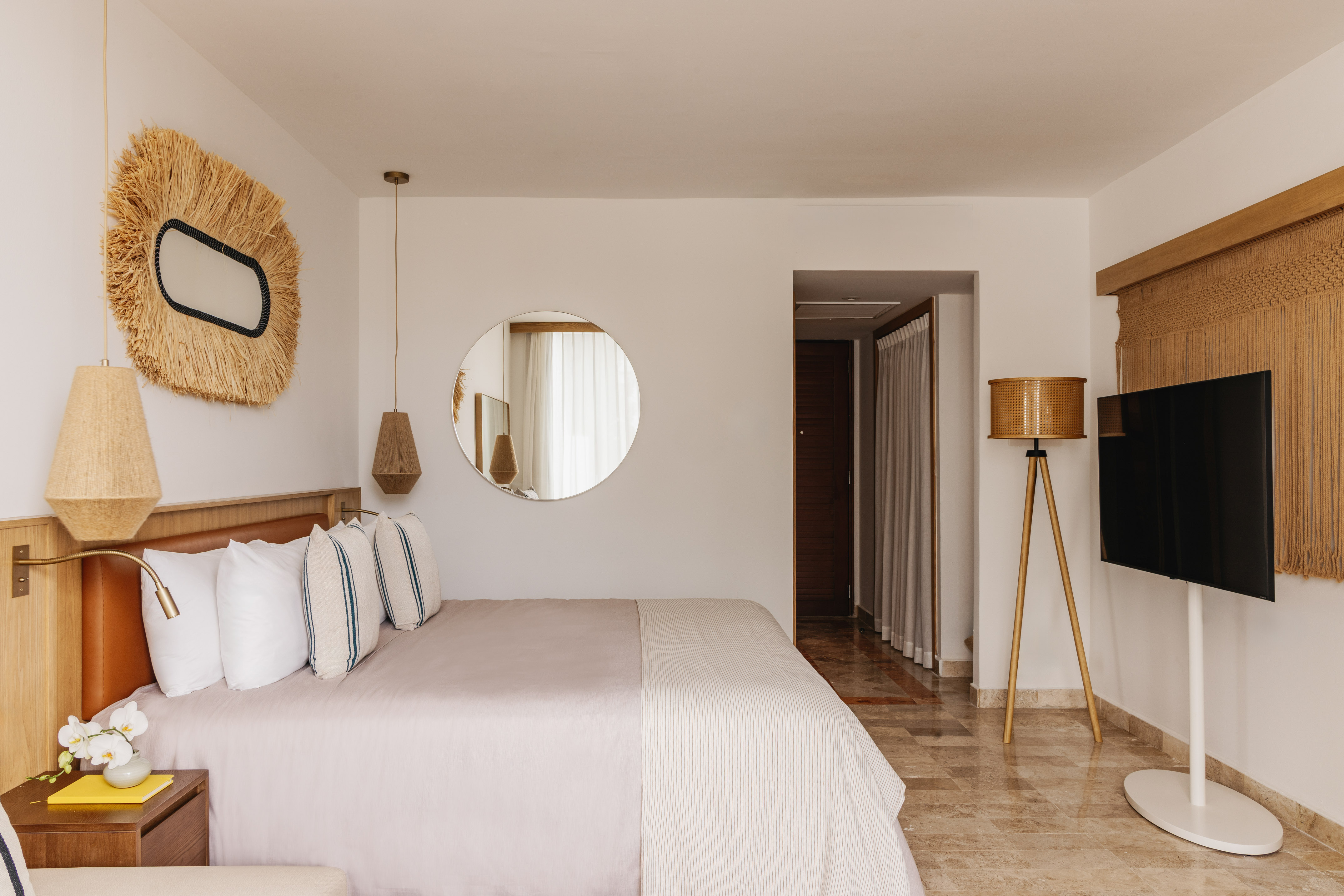 Modern boho hotel room with large bed, straw art, round mirror, and TV.